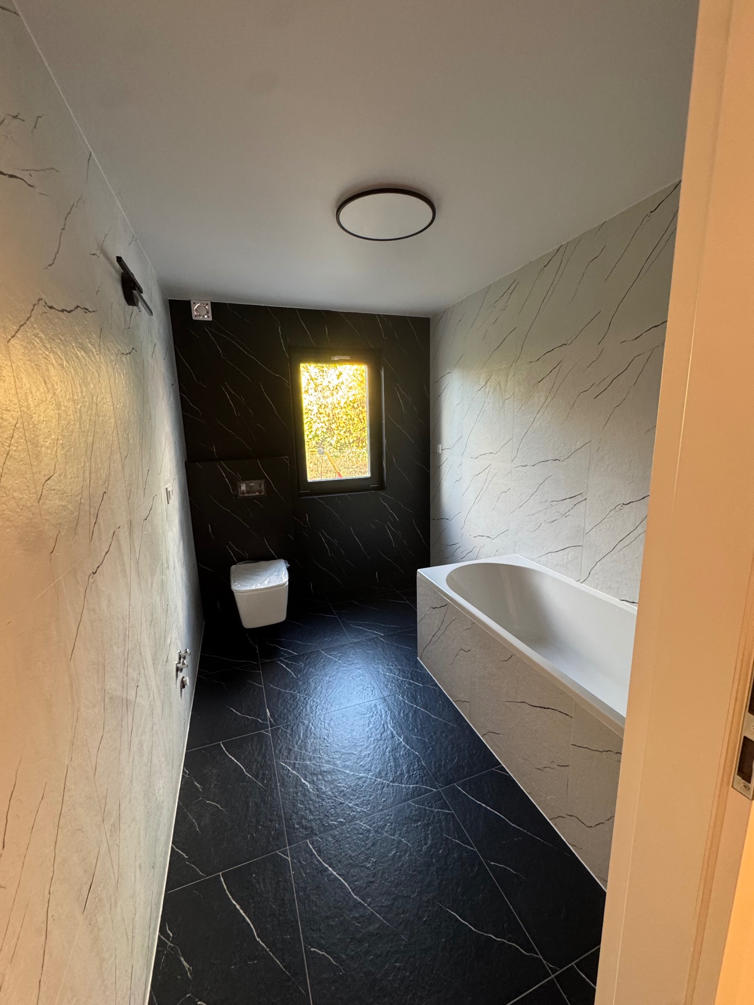 Nowoczesna łazienka z czarno-białymi płytkami imitującymi marmur, białą wanną i toaletą. Okno z widokiem na zieleń. Minimalistyczne oświetlenie sufitowe.