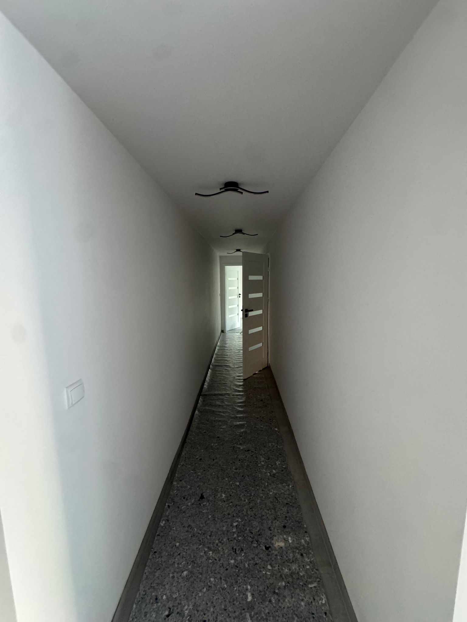 Biały korytarz z otwartymi drzwiami na końcu, zabezpieczona podłoga, minimalistyczne lampy sufitowe. Widok w perspektywie z niskiego kąta.