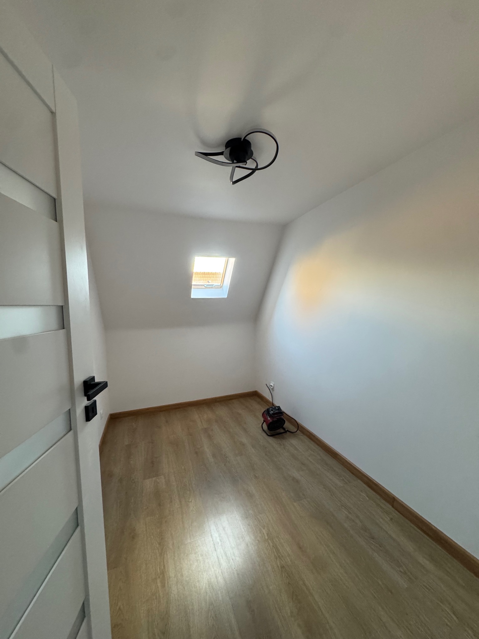 Poddasze z oknem dachowym i panelami podłogowymi w odcieniach drewna. Widoczna lampa sufitowa i białe ściany. Drzwi z czarną klamką po lewej stronie kadru.