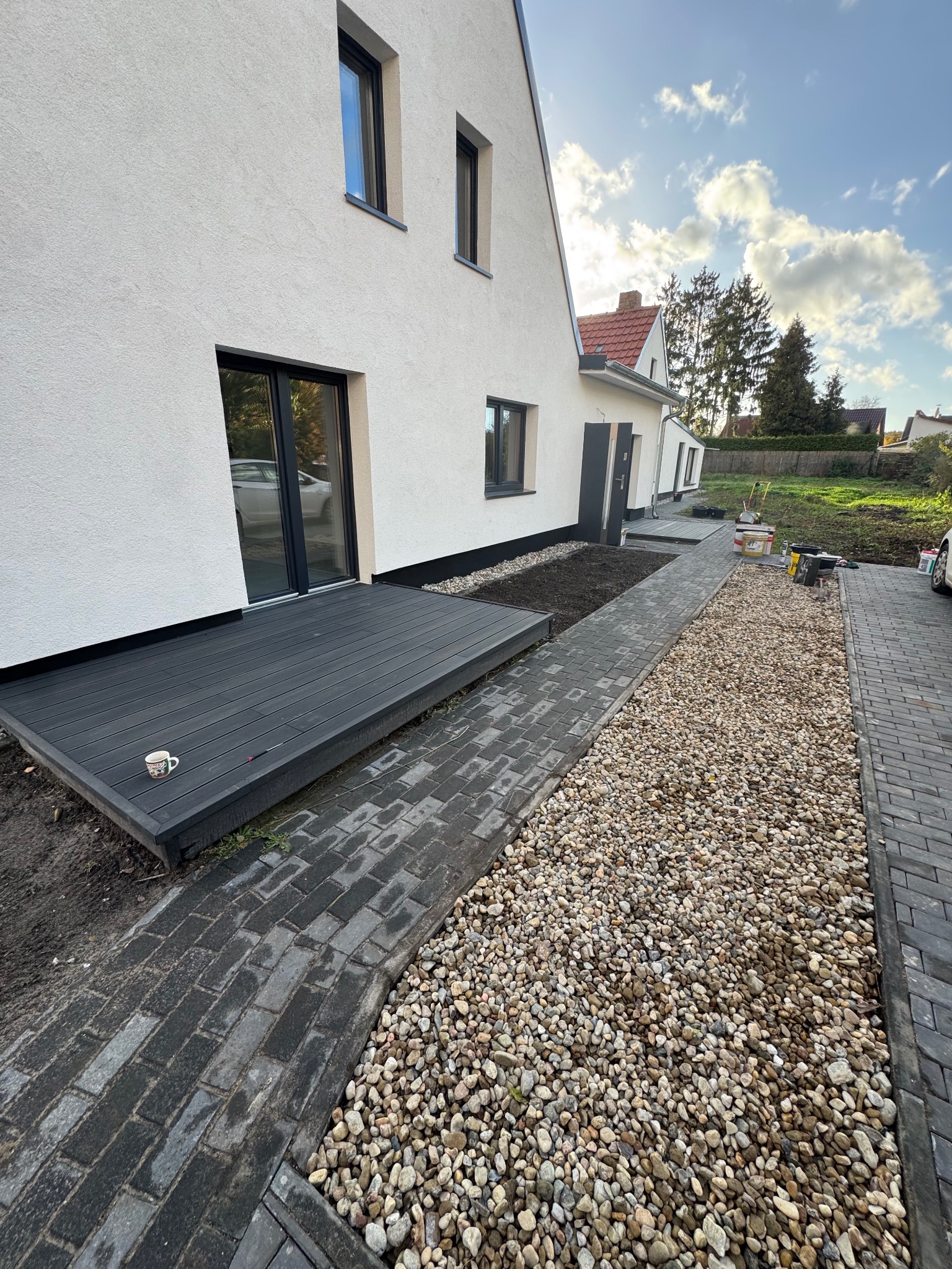 Nowoczesny taras z desek kompozytowych w kolorze grafitowym przylegający do białego domu z czarnymi oknami. Ścieżka z kostki brukowej i kamieni ozdobnych.
