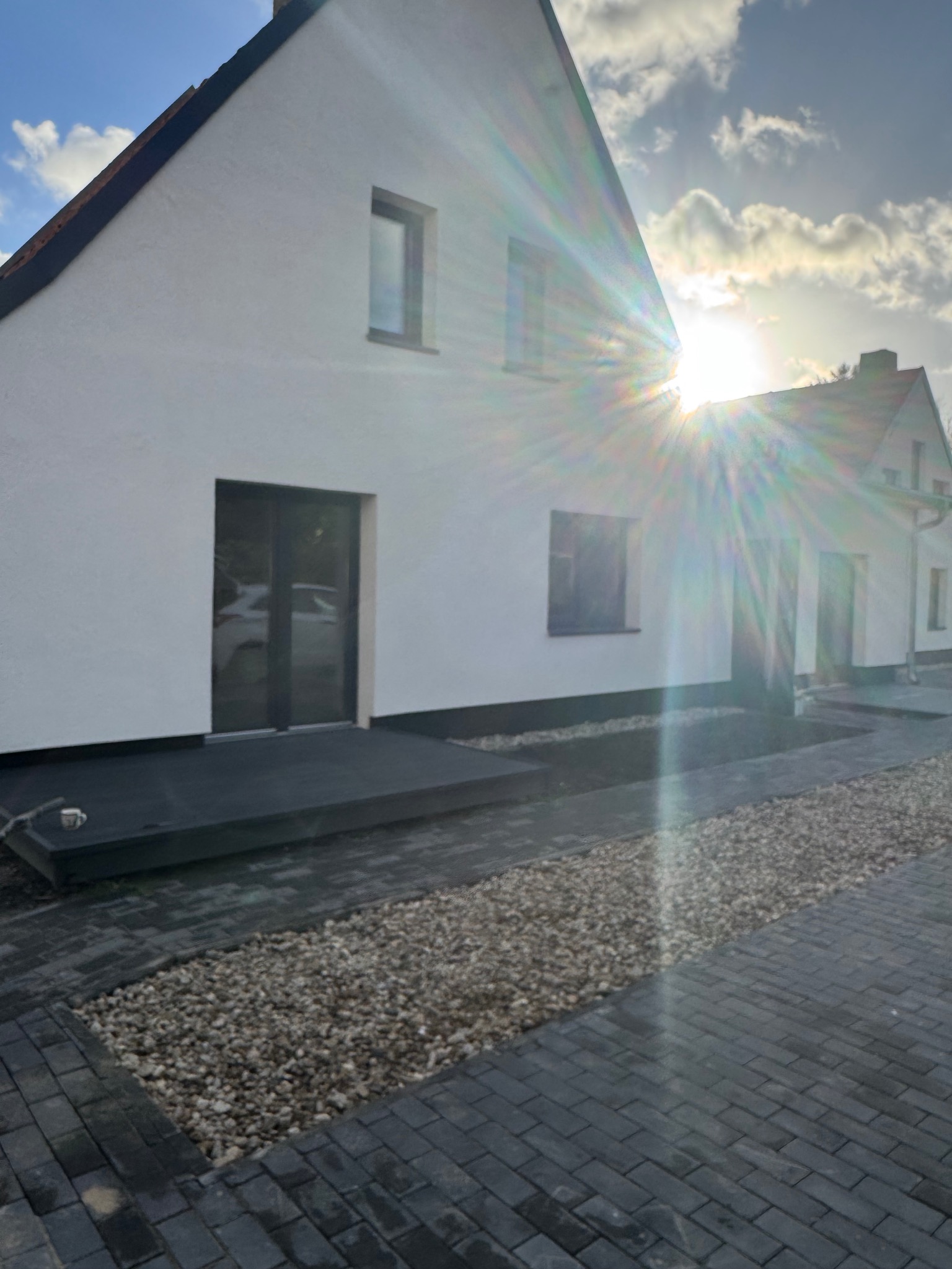 Biały dom z ciemną stolarką okienną i drzwiami, widok z zewnątrz, z brukowanym podjazdem i kamiennym rabatem. Słońce tworzy efekt rozbłysku.