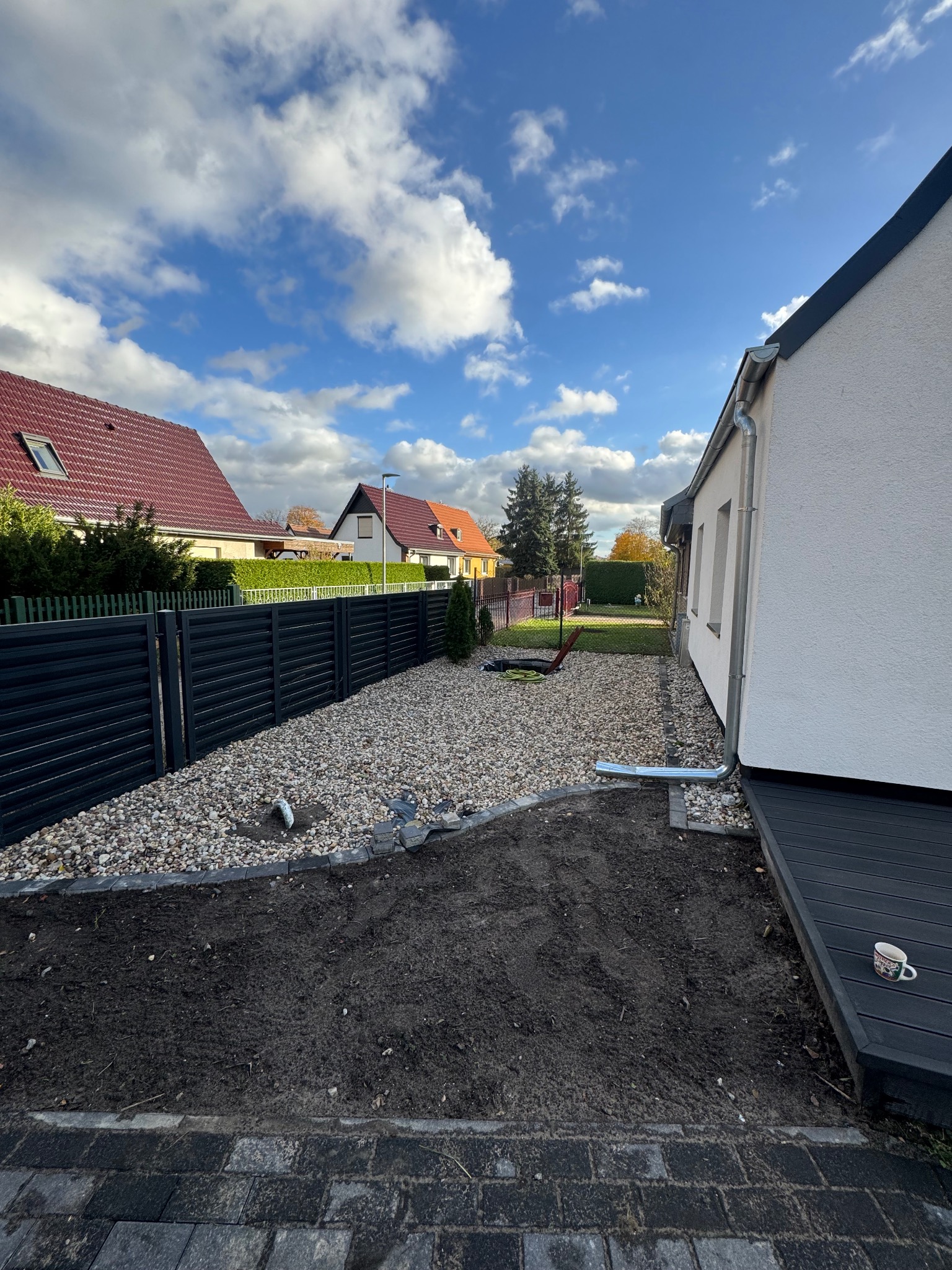 Nowoczesne ogrodzenie panelowe w kolorze antracytu oddziela posesję. Część działki wysypana drobnymi kamieniami, przygotowany teren pod trawnik. Widok na domy w tle.