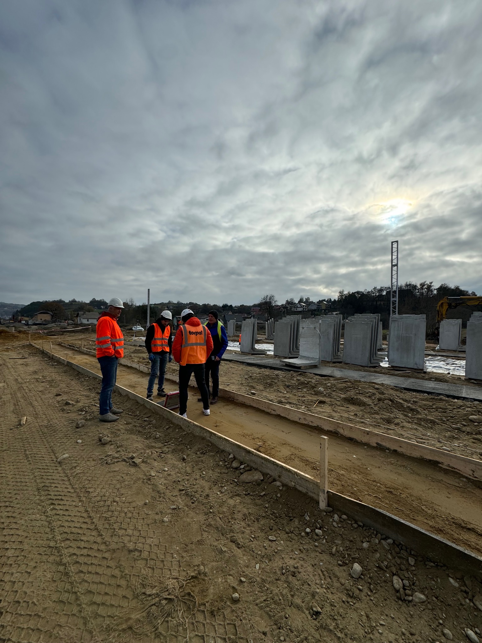 Ekipa budowlana w pomarańczowych kamizelkach omawia montaż prefabrykowanych elementów betonowych na placu budowy w pochmurny dzień. Widoczne szalunki.
