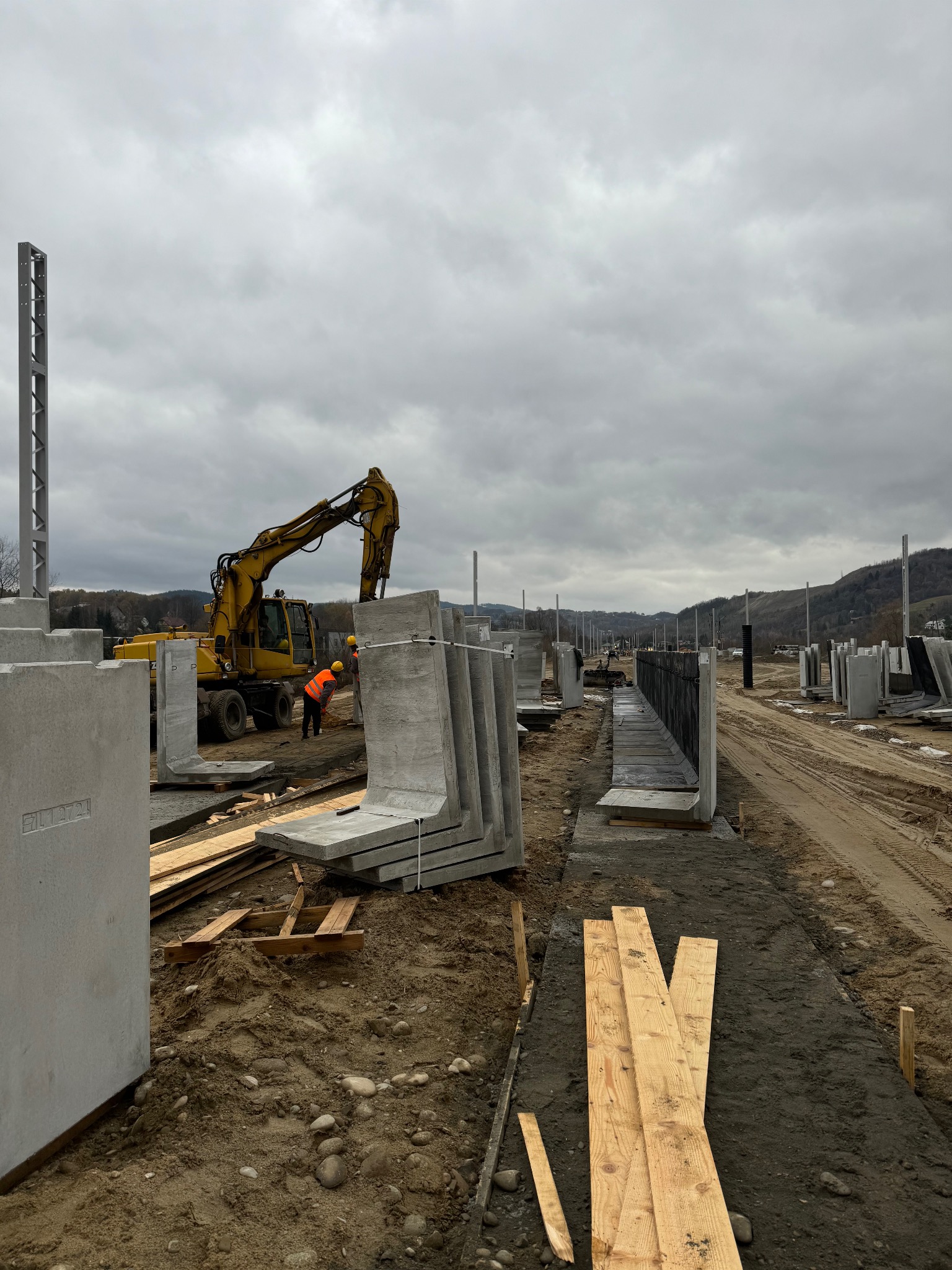 Montaż prefabrykowanych fundamentów z użyciem koparki na placu budowy. Widoczne elementy fundamentów typu L i pracownicy w odblaskowych kamizelkach.