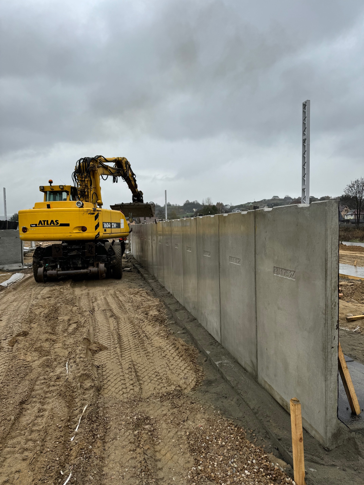Żółta koparka Atlas ustawia prefabrykowane betonowe panele ogrodzeniowe z wytłoczonymi numerami na placu budowy pod pochmurnym niebem.