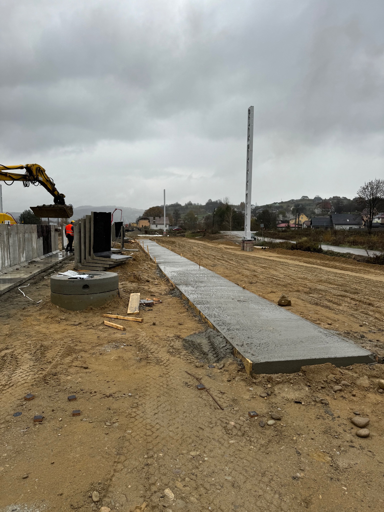 Wykop z betonową wylewką w szalunku, zbrojone prefabrykaty i koparka w tle, teren budowy w pochmurny dzień.