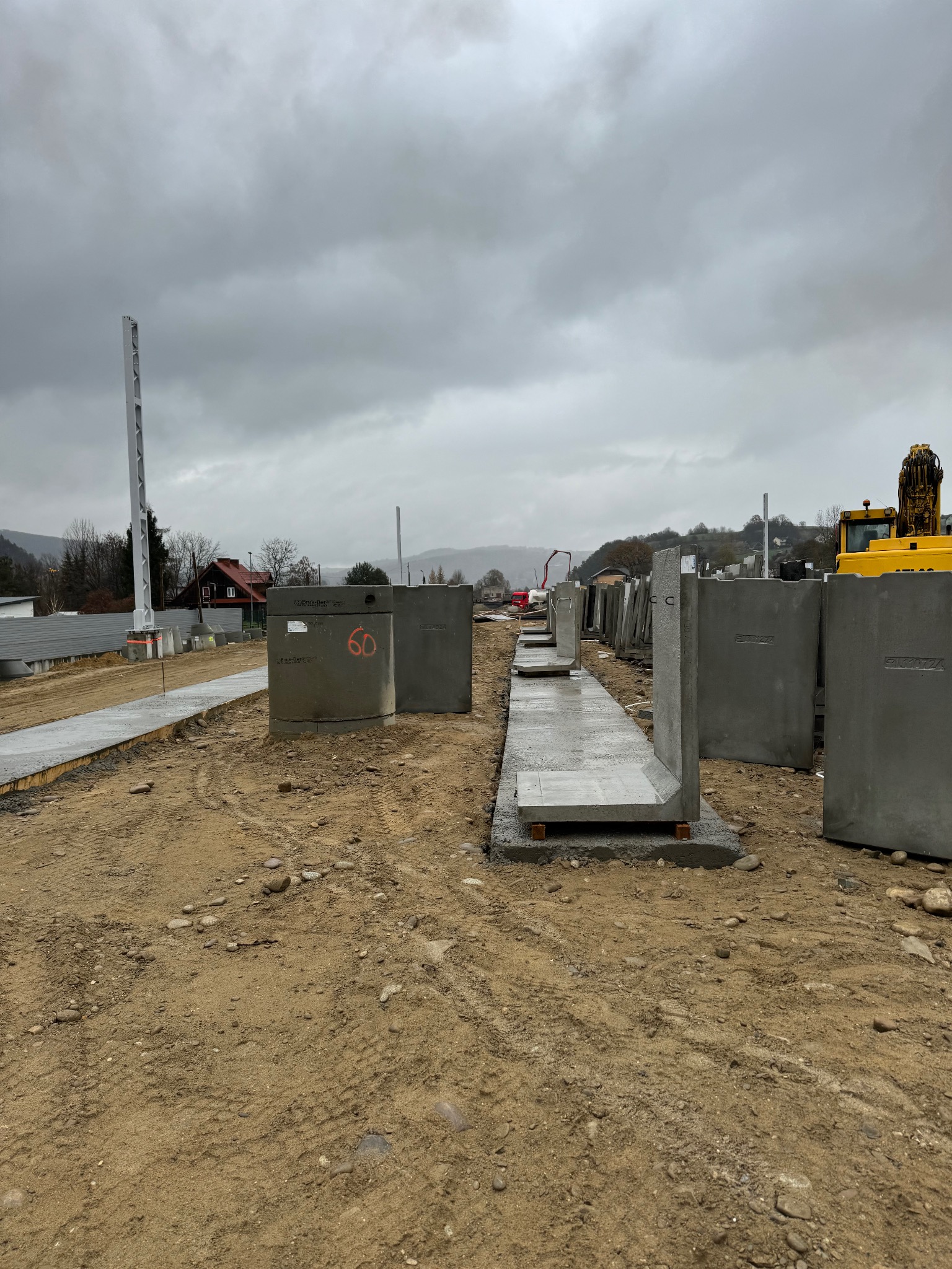 Betonowe prefabrykaty fundamentowe ułożone na placu budowy, widoczne elementy szalunków i ciężki sprzęt w tle, pochmurne niebo.