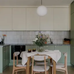 Jasna kuchnia z zielonymi szafkami i szarymi płytkami, st&oacute;ł z bukietem kwiat&oacute;w, oświetlona witryna z książkami i szkłem.