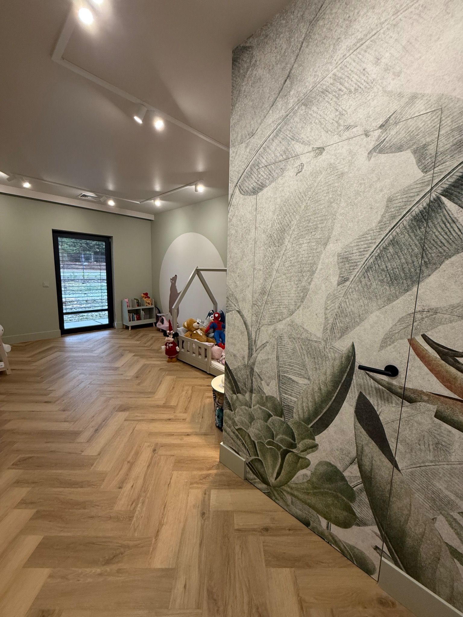 Pokój dziecięcy z drewnianą podłogą ułożoną w jodełkę, łóżkiem w kształcie domku, ścianą z motywem liści tropikalnych i okrągłym malunkiem na ścianie w tle.