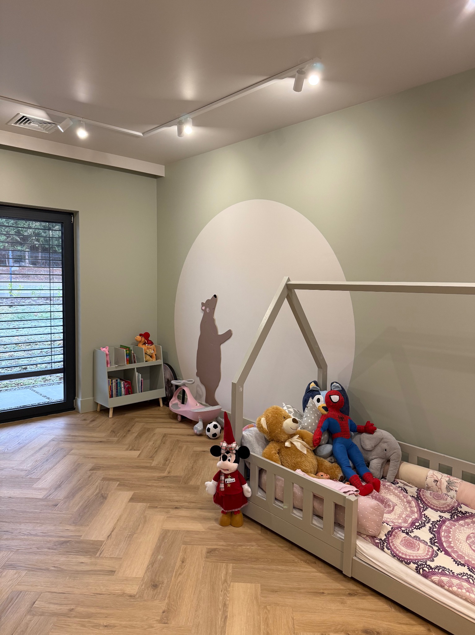 Jasny pokój dziecięcy z łóżkiem w kształcie domku, pomalowaną ścianą z motywem niedźwiedzia, drewnianą podłogą w jodełkę i zabawkami, oświetlony punktowymi lampami sufitowymi.