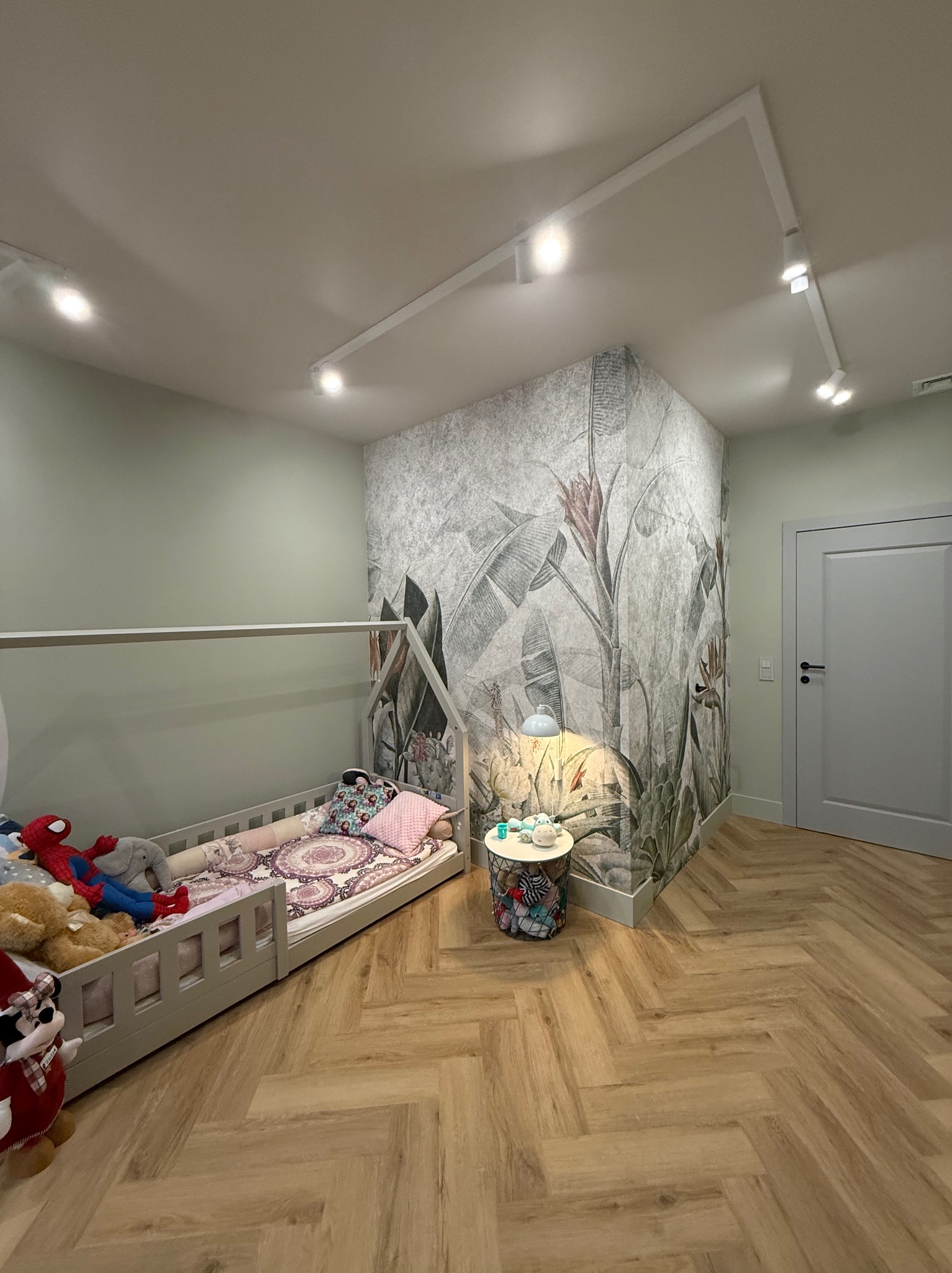 Pokój dziecięcy z łóżkiem w kształcie domku, fototapetą z motywem dżungli i parkietem ułożonym w jodełkę, oświetlony reflektorami na szynoprzewodzie.