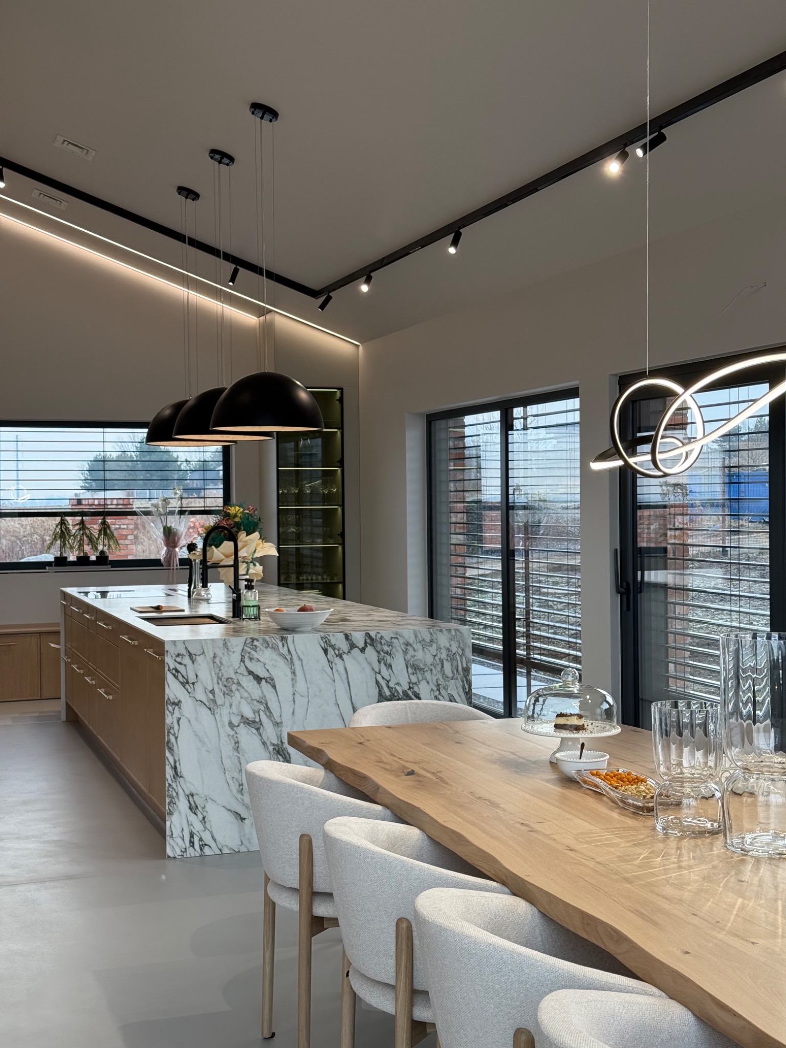 Nowoczesna kuchnia z wyspą wykończoną marmurem, drewnianym stołem z krzesłami i designerskim oświetleniem sufitowym.