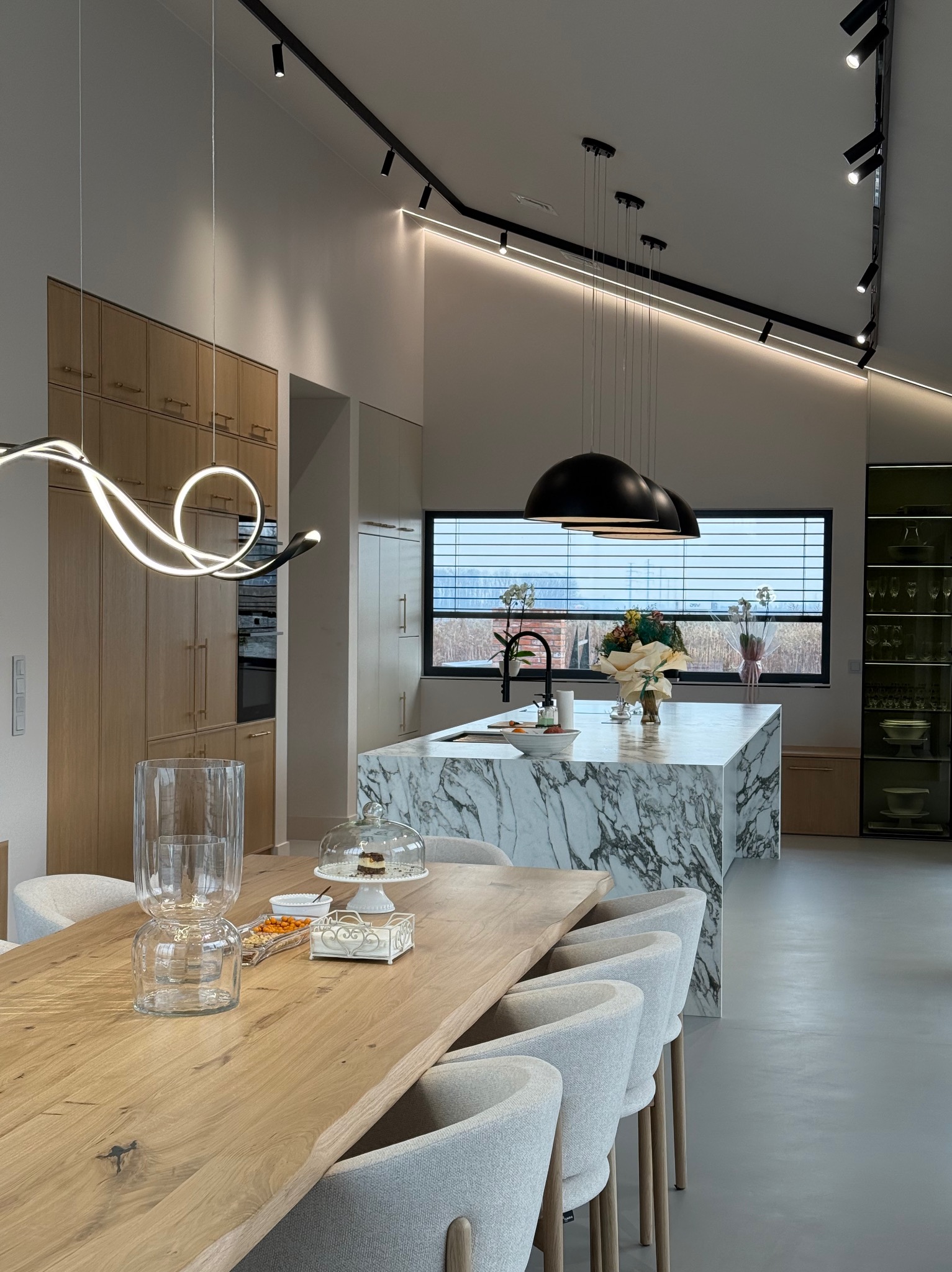 Nowoczesna kuchnia z marmurową wyspą, drewnianym stołem i designerskim oświetleniem, widok na okno z żaluzjami.