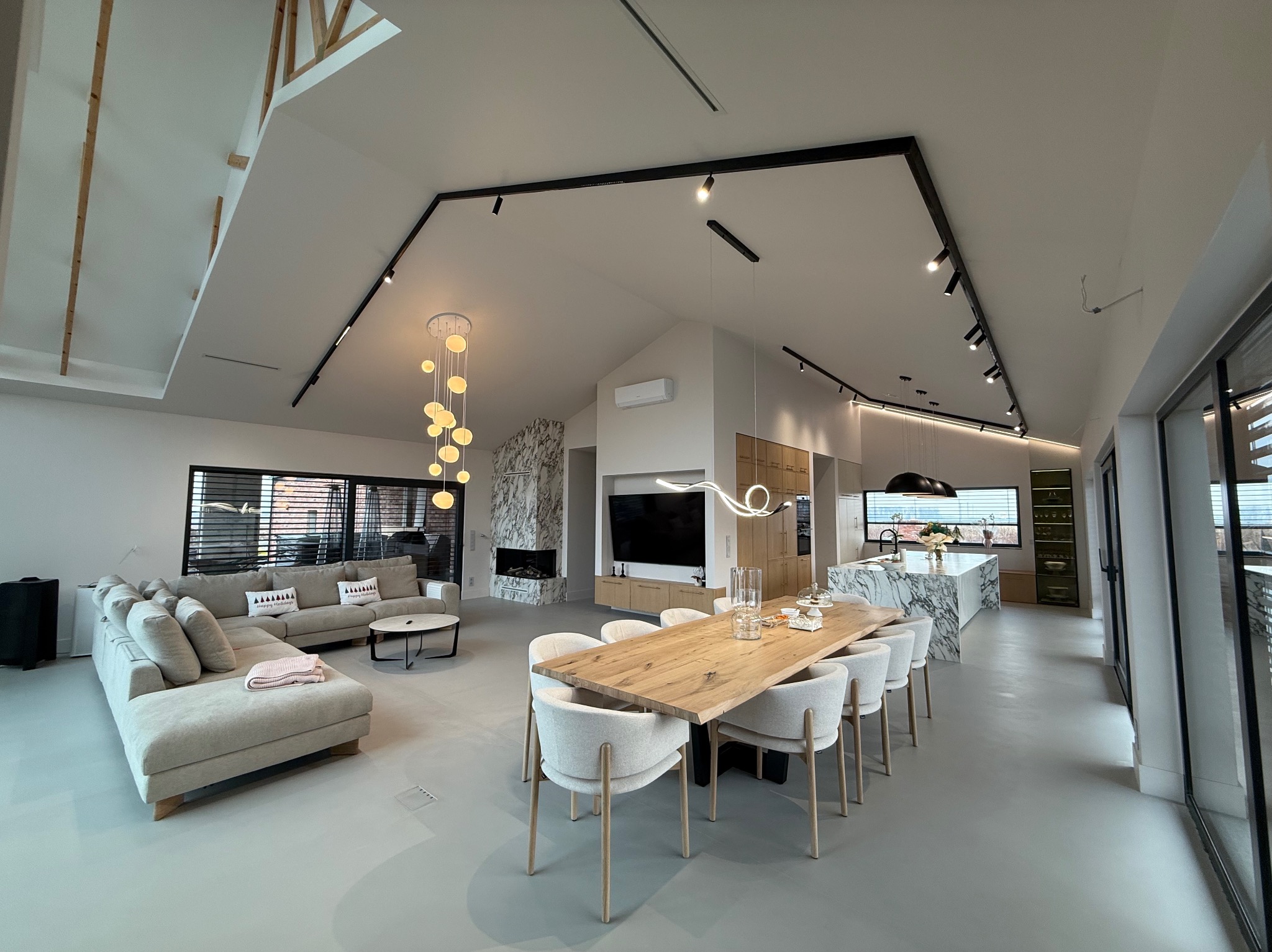 Przestronne wnętrze domu z szarym betonowym wykończeniem podłogi, oświetleniem szynowym na suficie i dużym drewnianym stołem jadalnym z krzesłami tapicerowanymi tkaniną boucle.