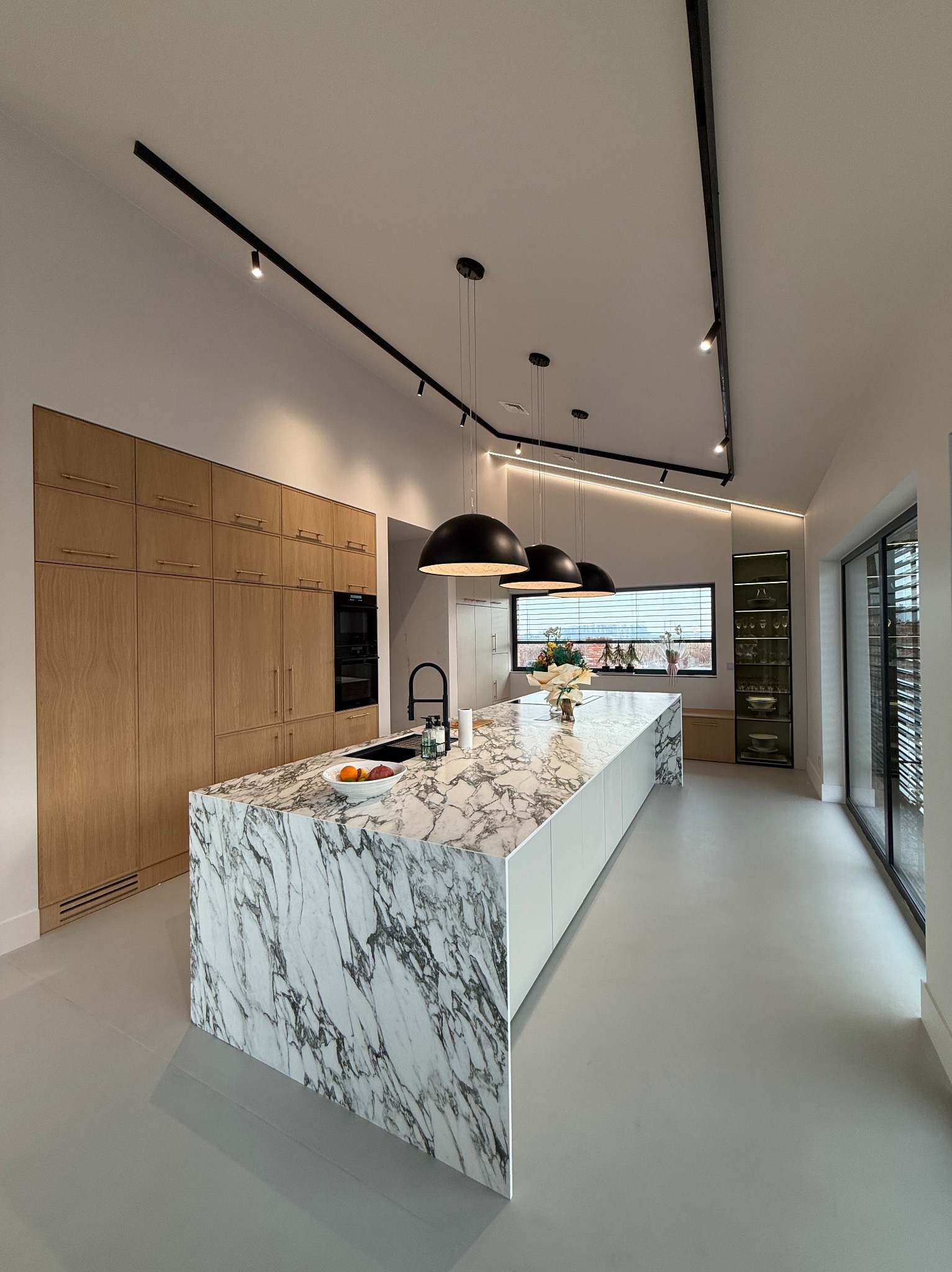 Nowoczesna kuchnia z marmurową wyspą, drewnianymi szafkami i czarnym oświetleniem szynowym, widok na okno z żaluzjami.