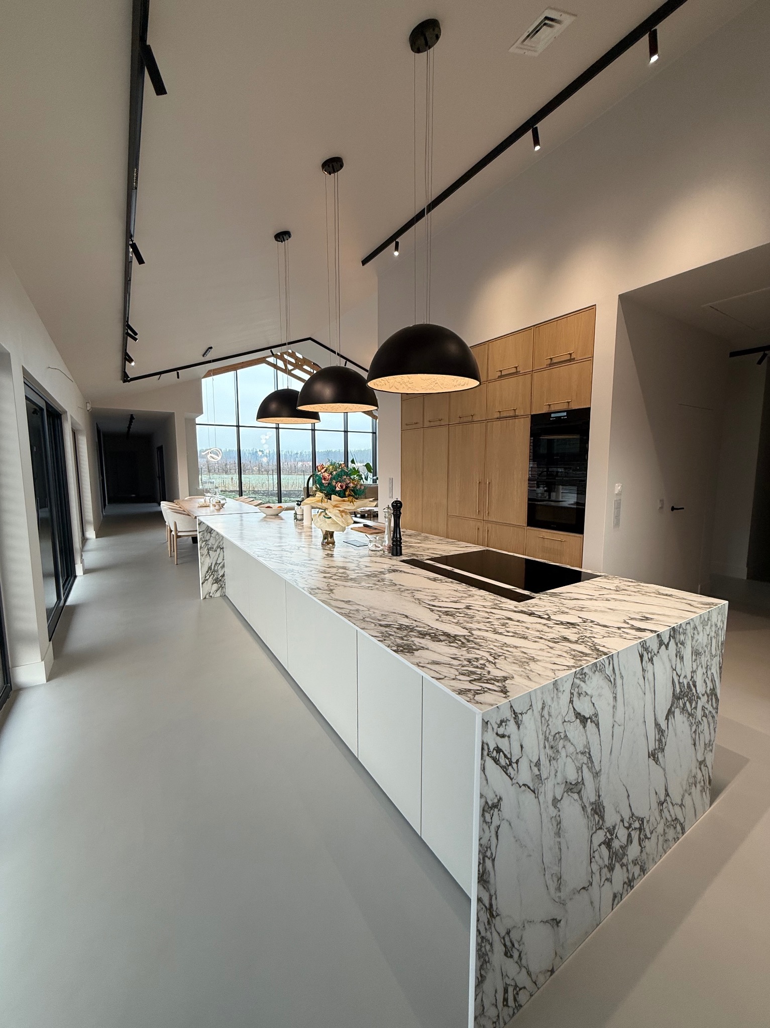 Przestronna kuchnia z wyspą o marmurowym blacie, minimalistycznymi białymi szafkami i wiszącymi lampami kopułowymi na tle jasnego wnętrza domu z dużymi oknami.
