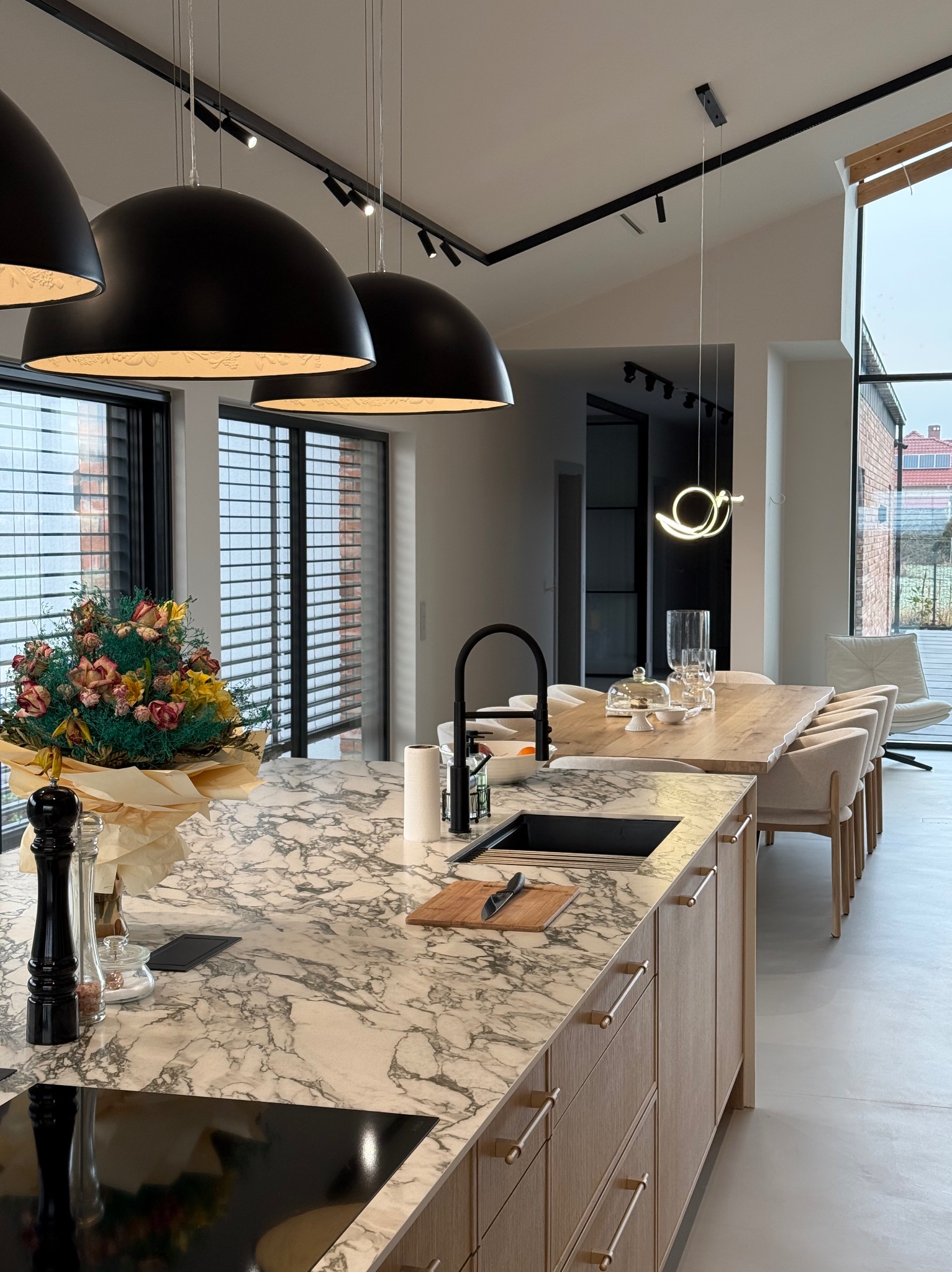 Nowoczesna kuchnia z wyspą z marmurowym blatem, wyposażona w czarny zlew i armaturę, drewniane szafki z poziomymi uchwytami, oraz bukiet kwiatów. Nad wyspą wiszą trzy duże, czarne lampy kopułowe.