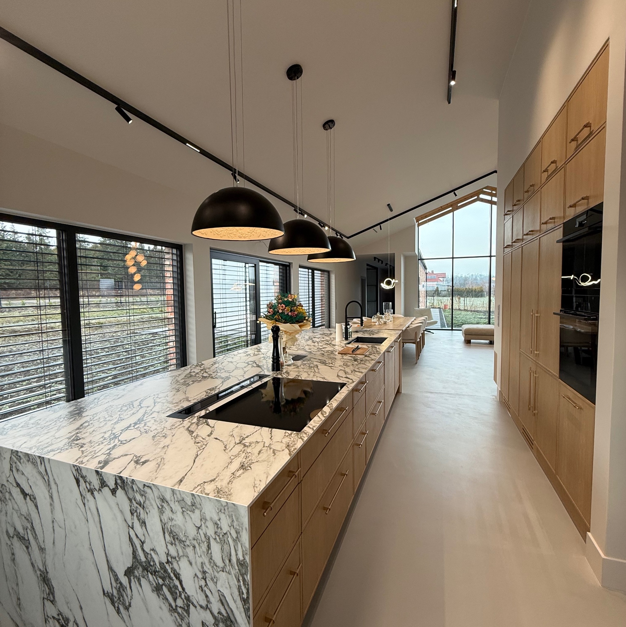 Nowoczesna kuchnia z marmurowym blatem wyspy, drewnianymi szafkami, czarnymi lampami wiszącymi i oknami z widokiem na ogród.