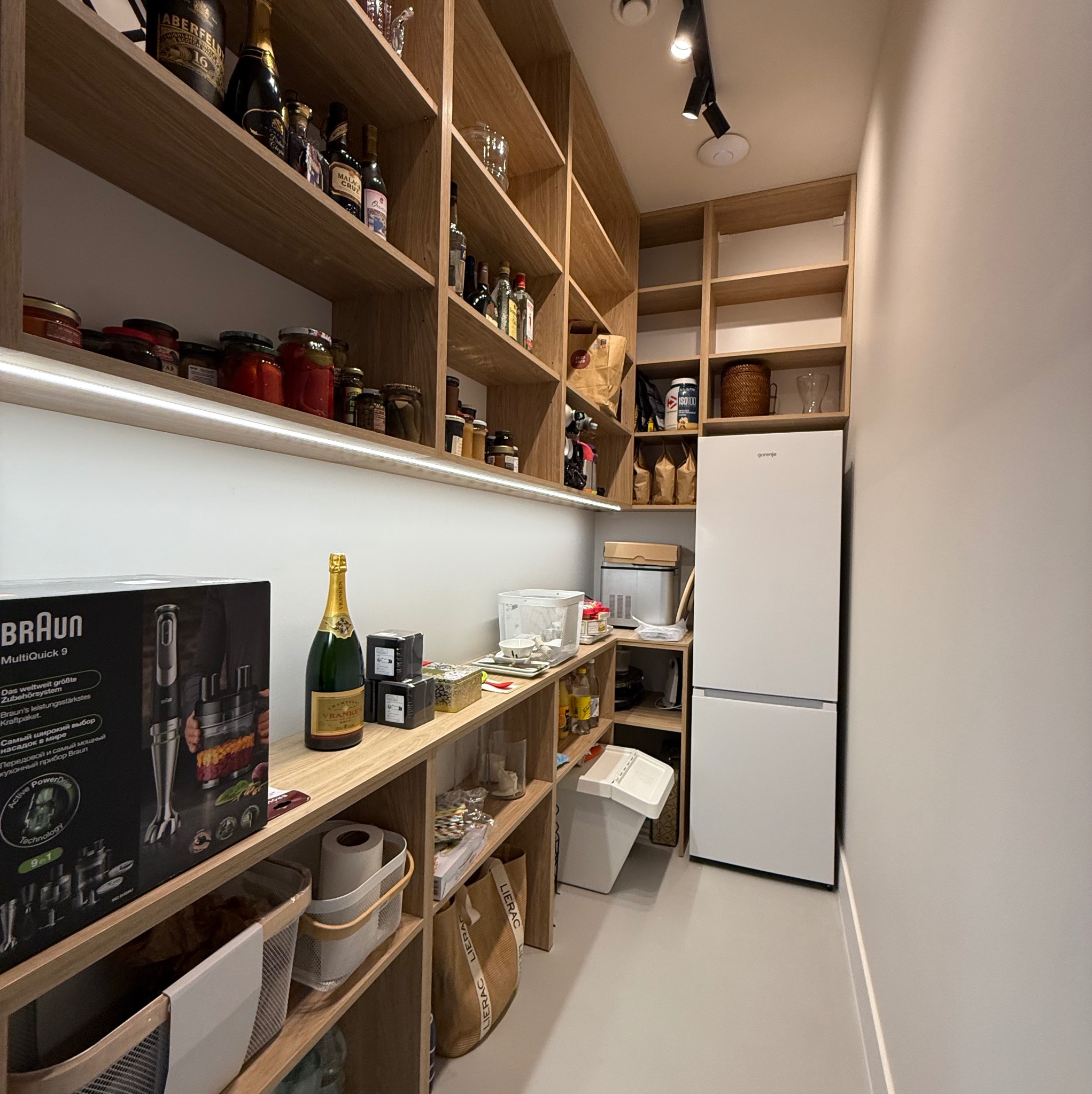 Spiżarnia z drewnianymi półkami wypełnionymi słoikami, butelkami, produktami spożywczymi, obok biała lodówka Gorenje, na półce stoi opakowanie blendera Braun MultiQuick 9 oraz butelka szampana...