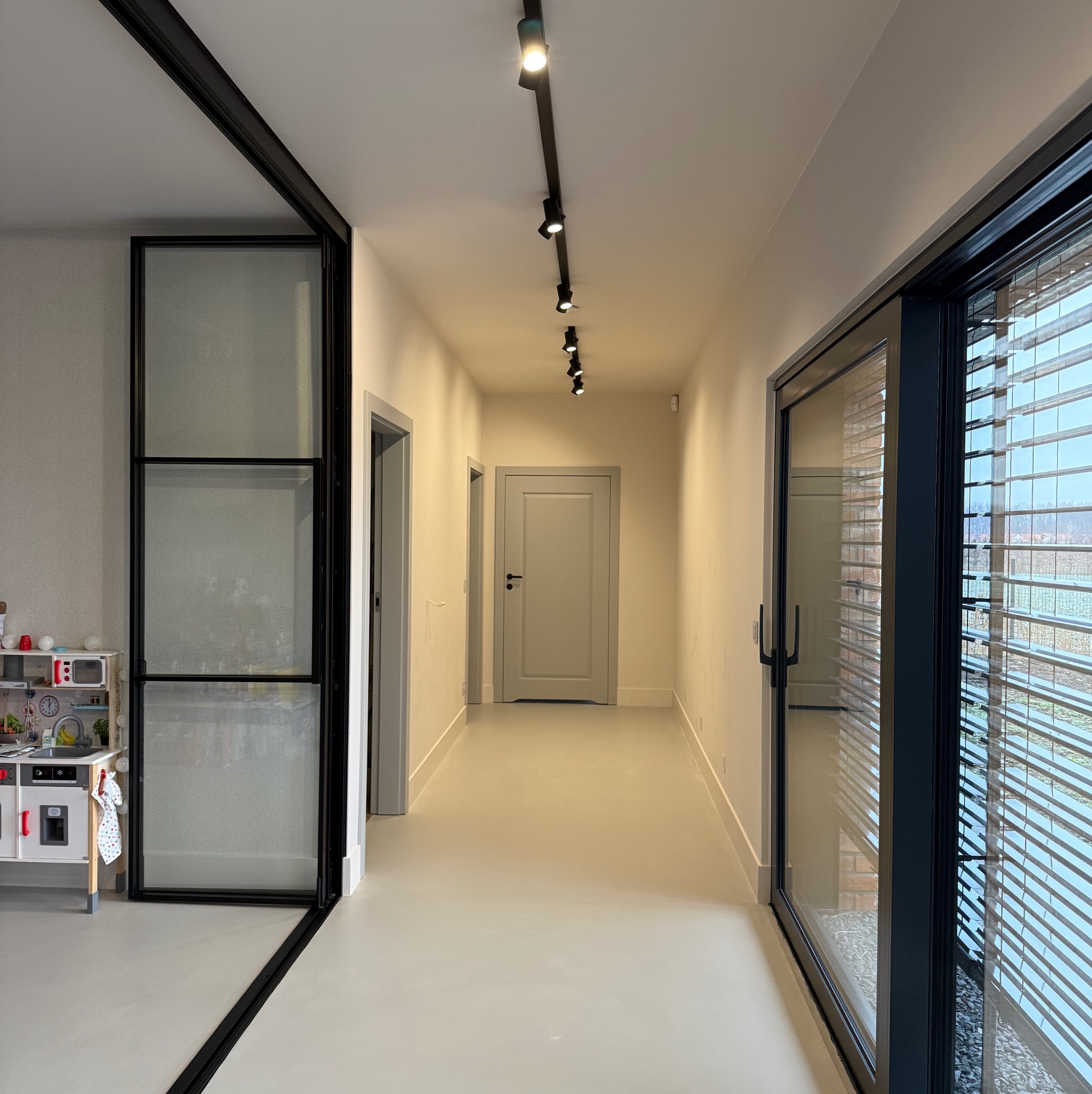 Nowoczesny korytarz z szarą podłogą żywiczną, oświetleniem szynowym, przeszklonymi drzwiami przesuwnymi w czarnej ramie i widokiem na zewnątrz przez żaluzje.