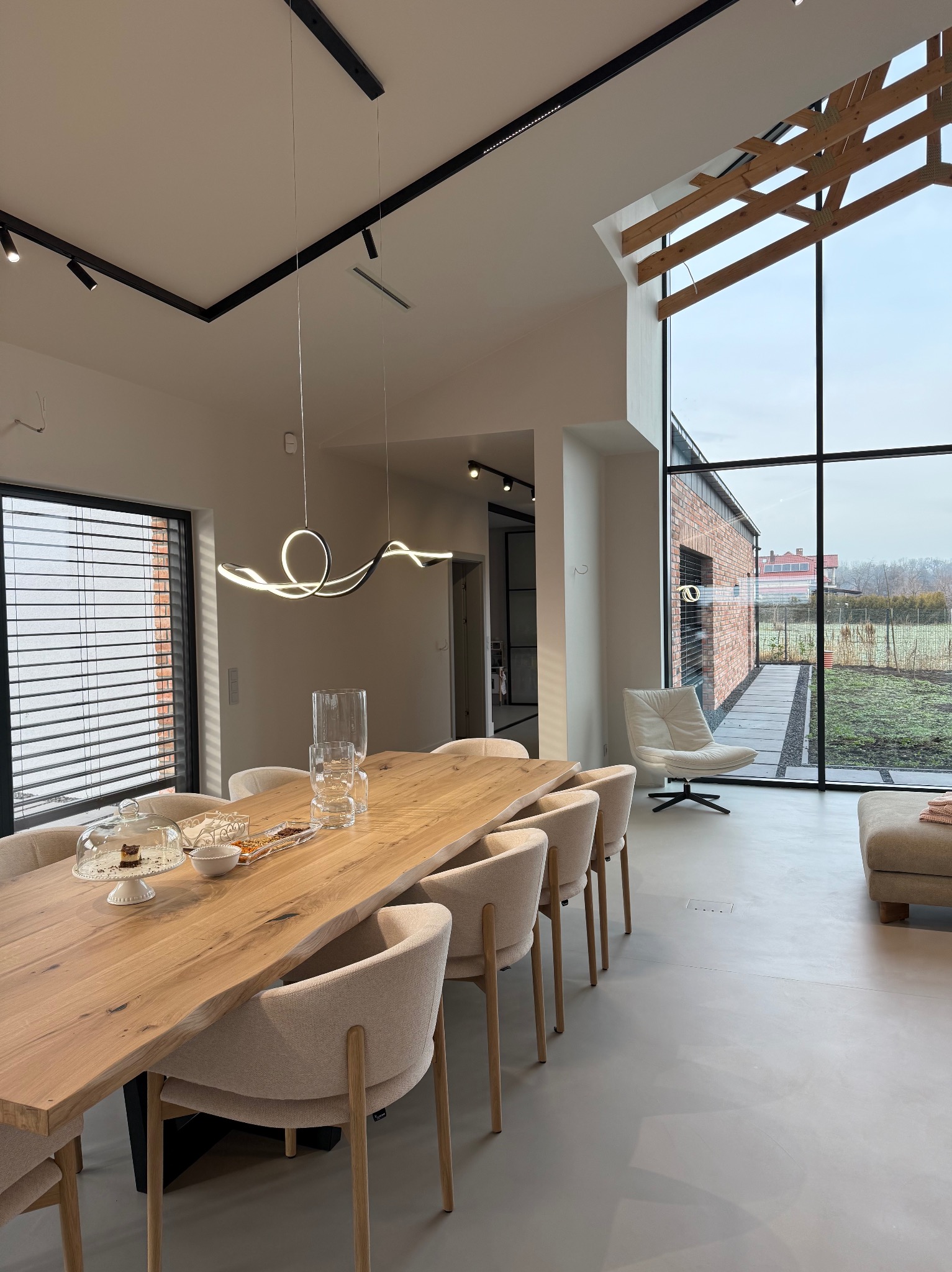 Jasne, przestronne wnętrze jadalni z dużym drewnianym stołem, krzesłami w kolorze ecru, nowoczesną lampą wiszącą i panoramicznym oknem z widokiem na ogród. Na stole szklana patera z ciastem...