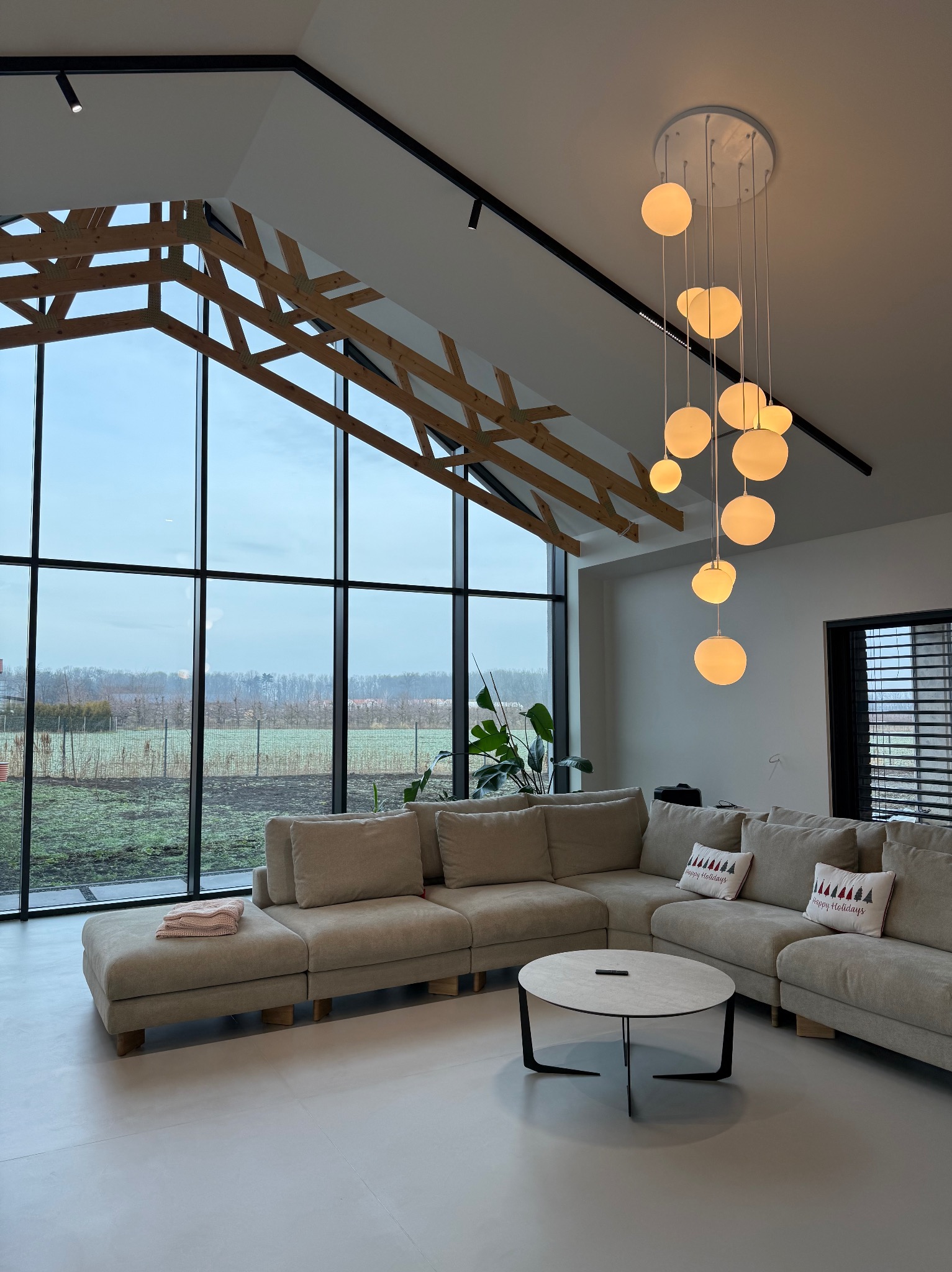Jasne wnętrze salonu z wysokim, przeszklonym oknem, drewnianą konstrukcją stropu i nowoczesną lampą wiszącą z kulistymi kloszami, beżowa sofa z poduszkami z motywem świątecznym, okrągły stolik kawowy.