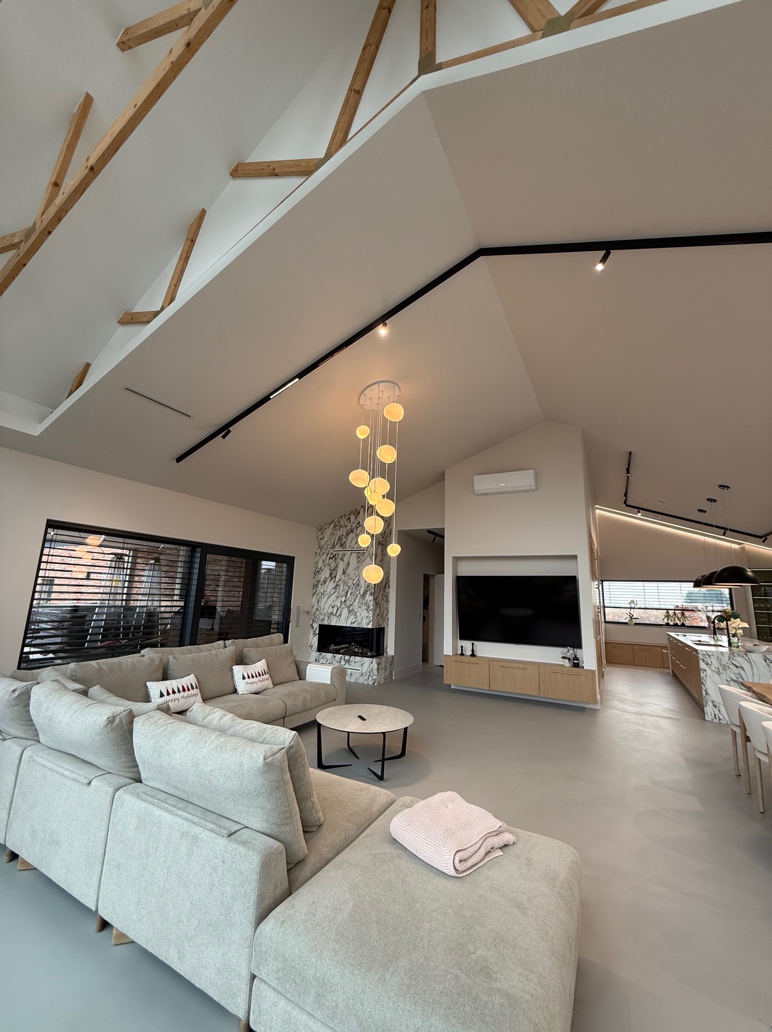 Przestronny salon z wysokim sufitem, drewnianymi belkami, szarą sofą modułową z poduszkami świątecznymi, marmurowym kominkiem i nowoczesnym oświetleniem.