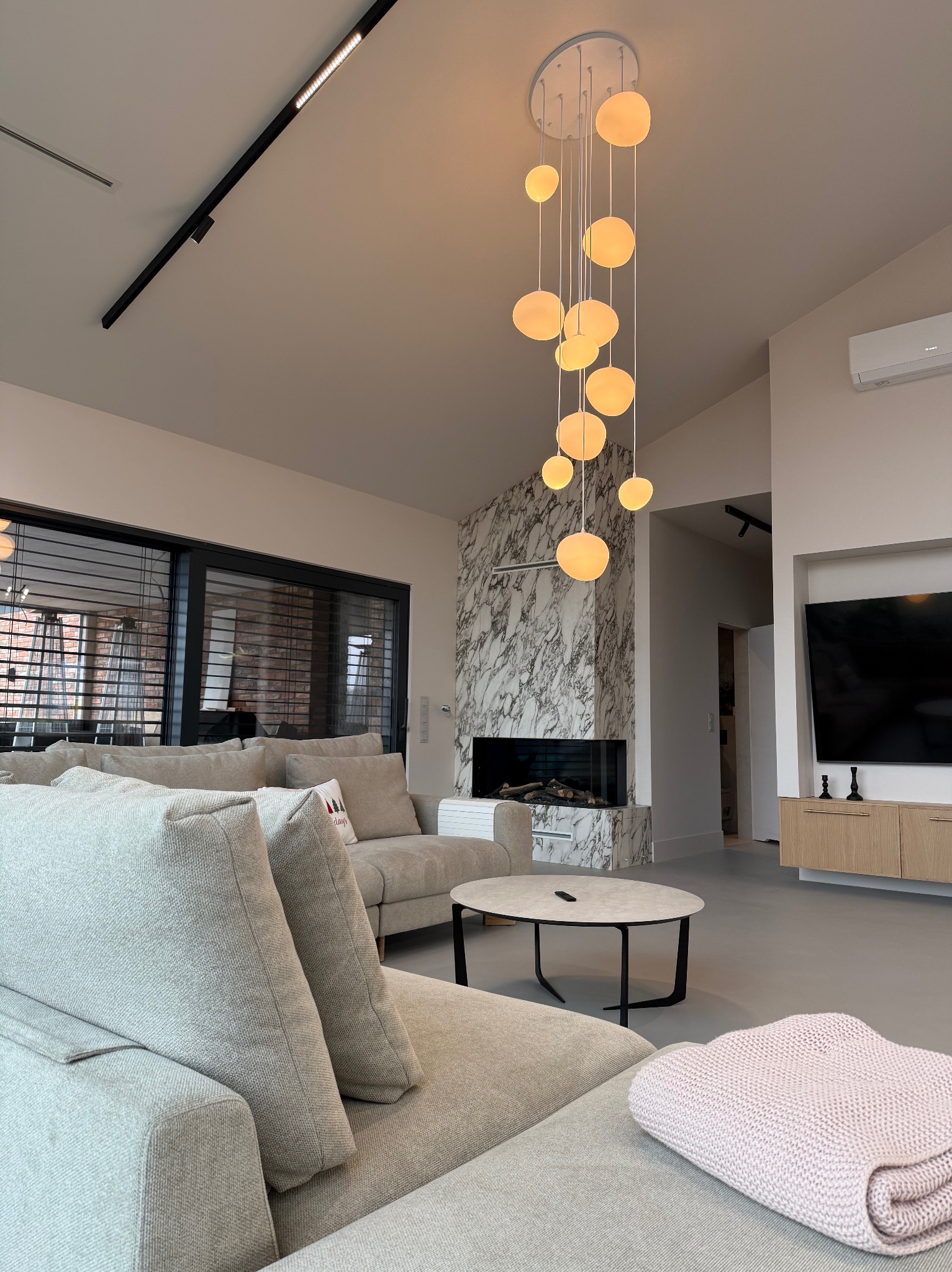 Nowoczesny salon z szarą sofą, marmurowym kominkiem, okrągłym stolikiem kawowym i wiszącą lampą z kulistymi kloszami, oświetlony światłem dziennym wpadającym przez okno z żaluzjami.