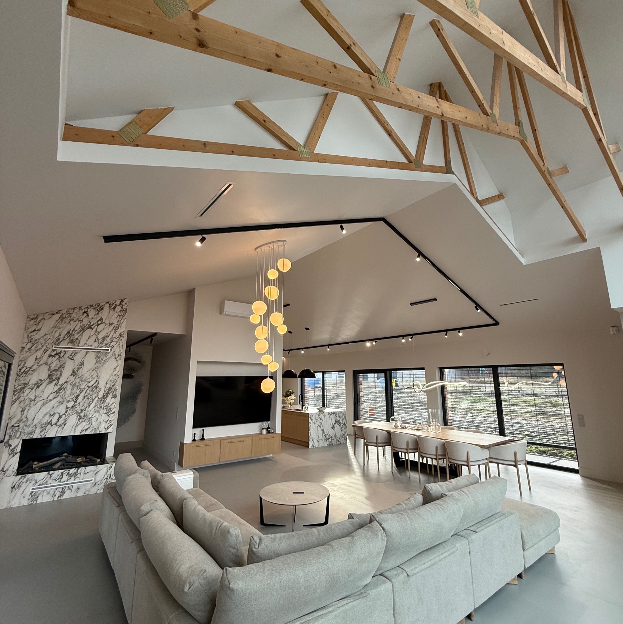 Przestronne wnętrze salonu z marmurowym kominkiem, drewnianymi belkami stropowymi i nowoczesnym oświetleniem, widok z perspektywy narożnika sofy.