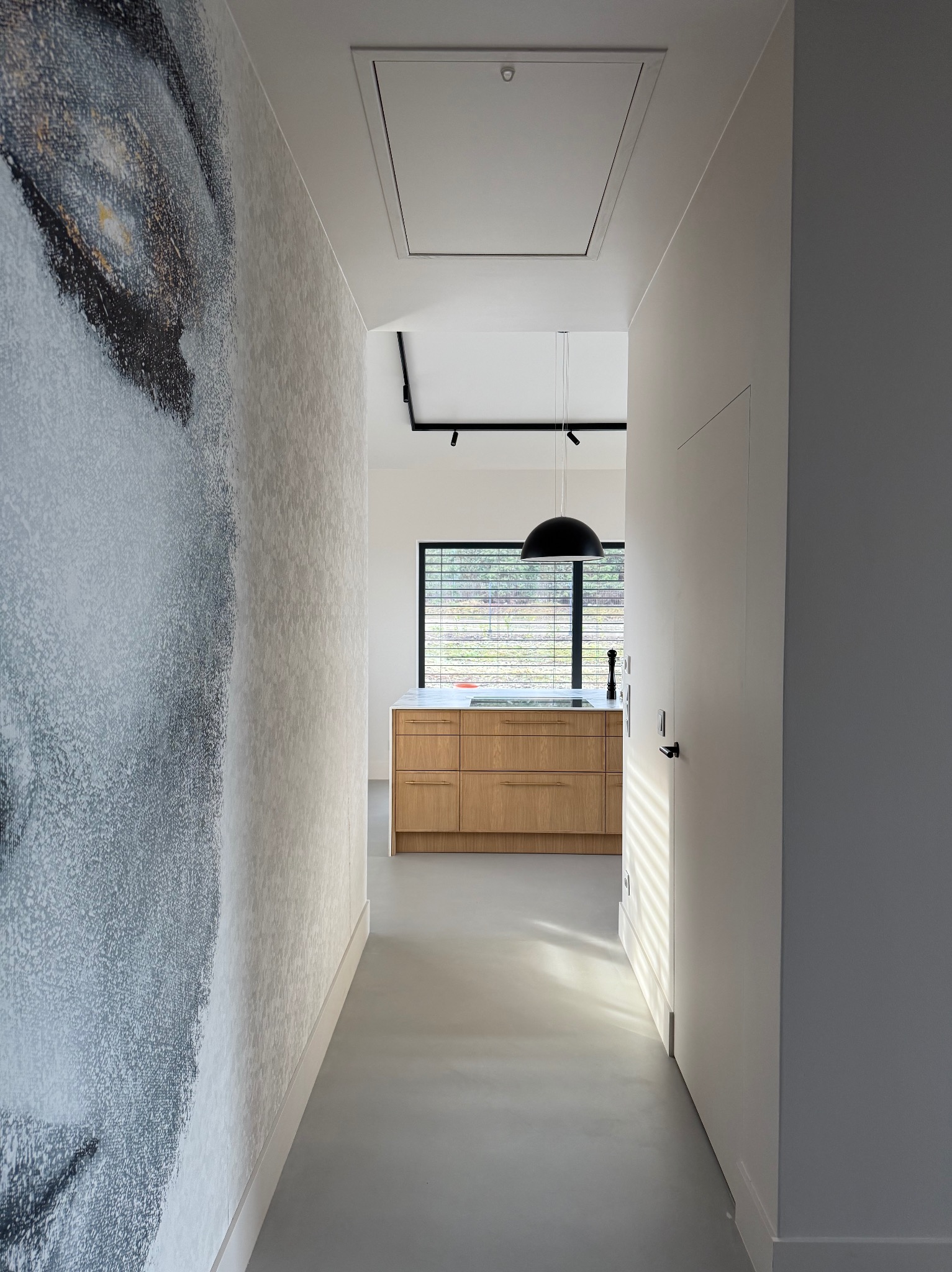Nowoczesny korytarz z betonową podłogą prowadzący do kuchni z drewnianymi szafkami, na ścianie abstrakcyjny mural, a w suficie klapa włazowa.