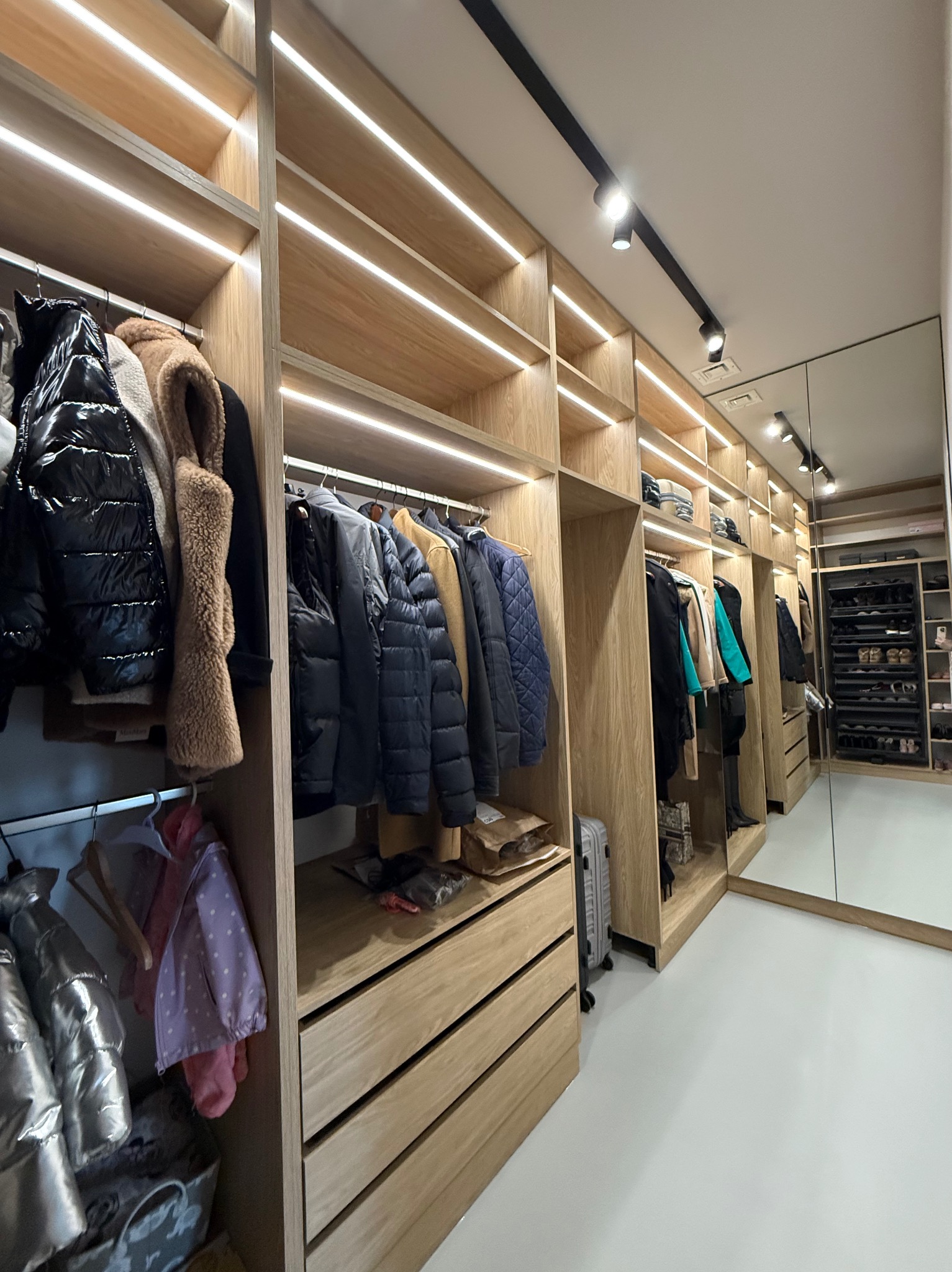 Przestronna garderoba z oświetleniem LED, drewnianymi półkami i szufladami, lustrzane drzwi ukazują dalszą część z półkami na buty.