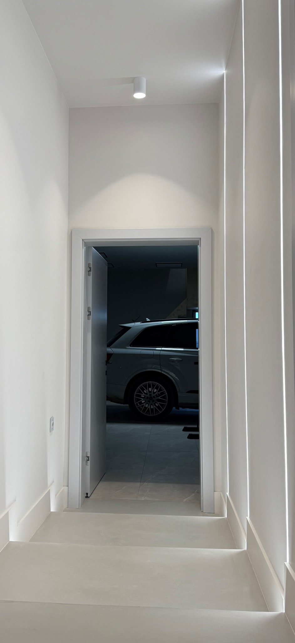 Nowoczesne, jasne wnętrze z minimalistycznym oświetleniem LED, widok na garaż z białym SUV-em przez otwarte drzwi.