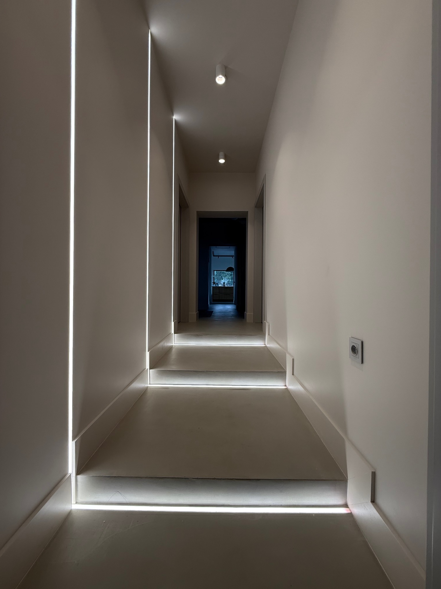 Nowoczesny korytarz z oświetleniem LED w ścianach i przypodłogowych listwach, wykończony w jasnych, neutralnych barwach, widoczny kontakt elektryczny na ścianie po prawej stronie.