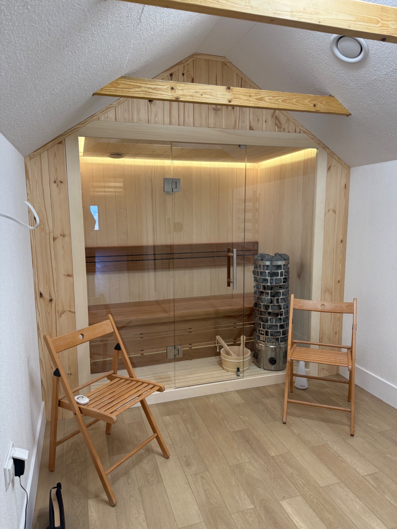 Wnętrze sauny w domowym zaciszu, z drewnianymi panelami, szklanymi drzwiami i piecem na kamienie, uzupełnione dwoma drewnianymi krzesłami.