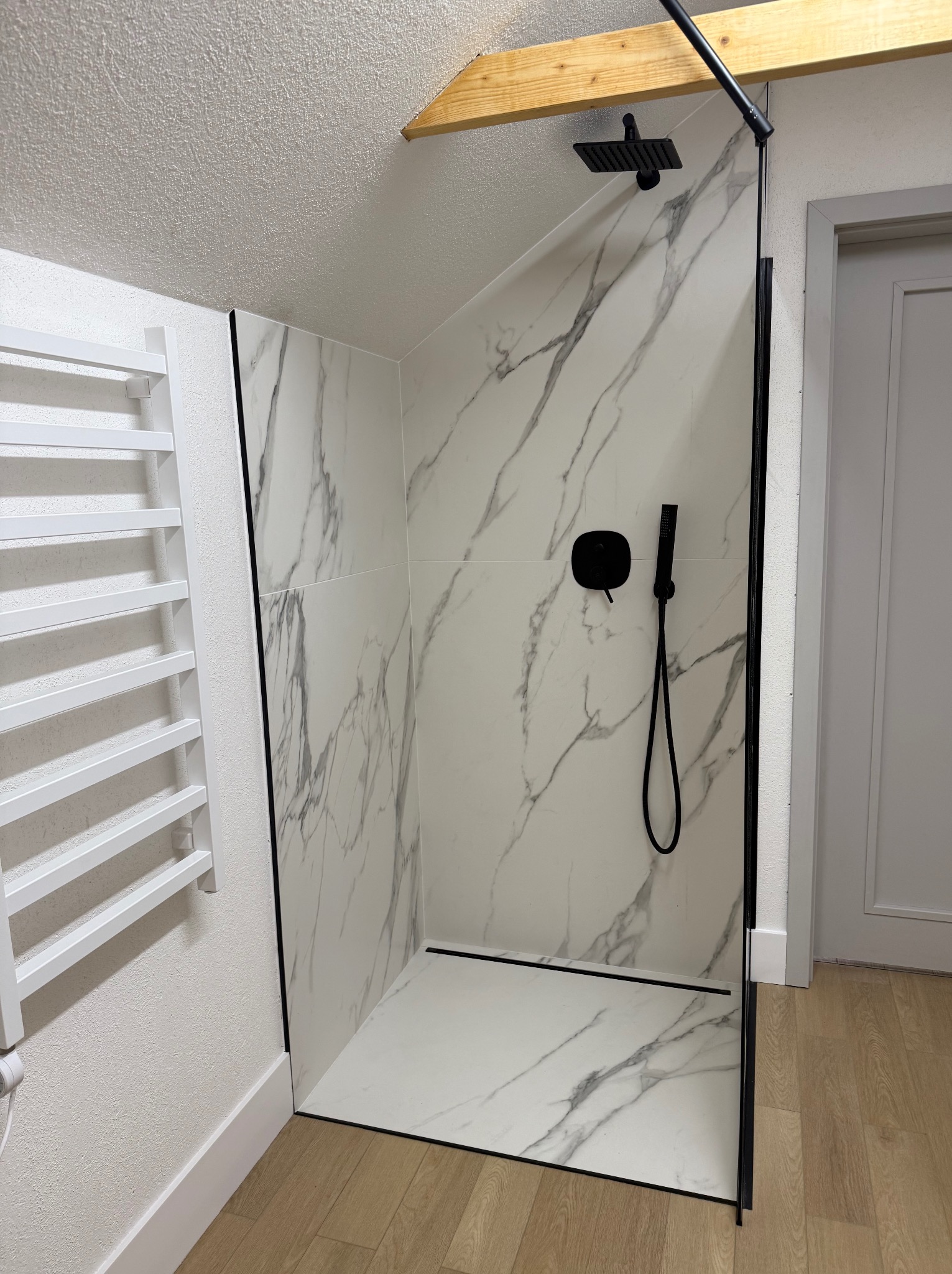 Nowoczesna kabina prysznicowa walk-in z białymi płytkami imitującymi marmur, czarną armaturą i grzejnikiem drabinkowym w wyremontowanej łazience na poddaszu z drewnianą belką stropową.