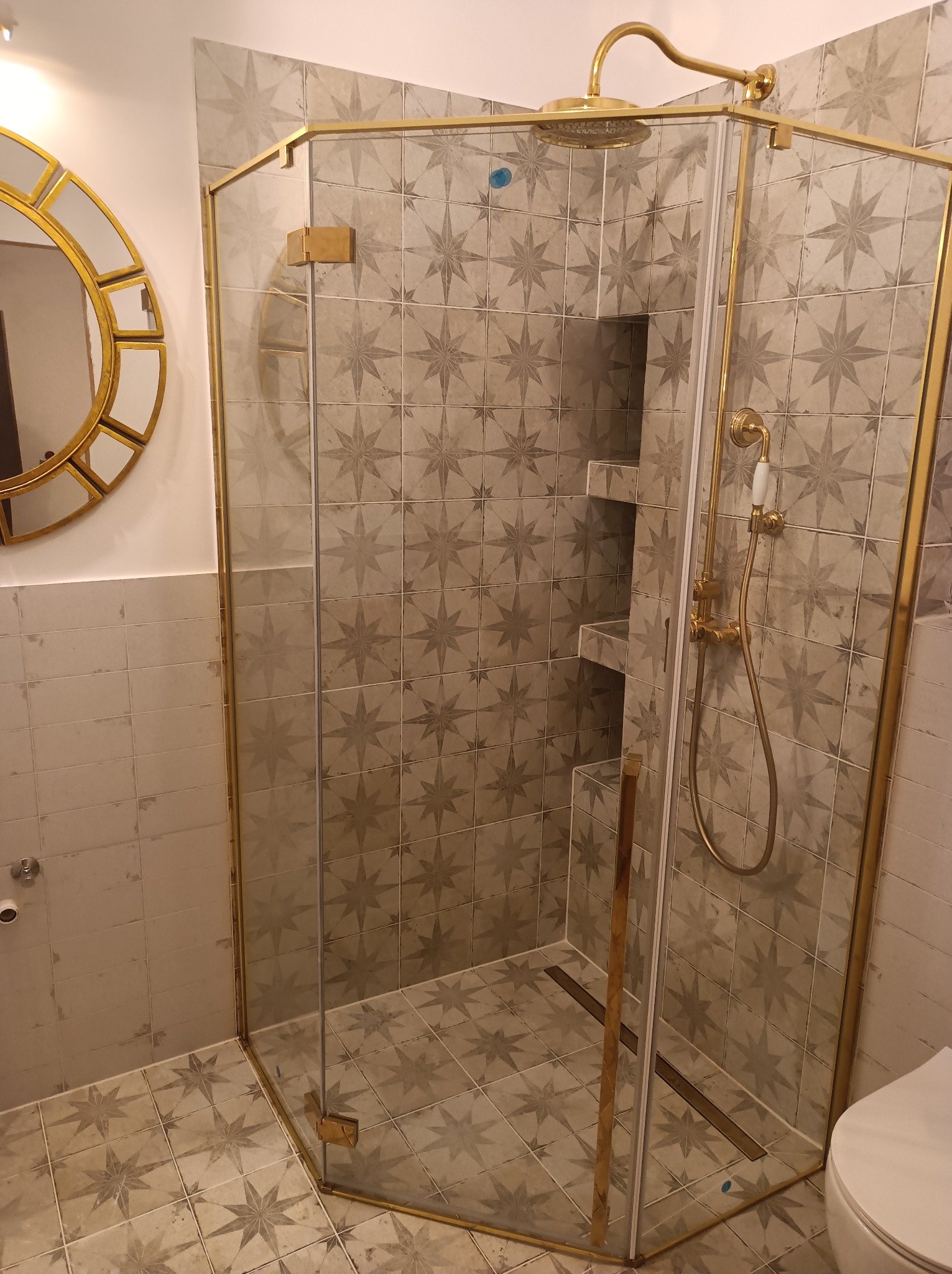 Kabina prysznicowa narożna z brodzikiem liniowym i złotymi profilami, płytki w gwiazdy na ścianach i podłodze, okrągłe lustro w złotej ramie w tle.