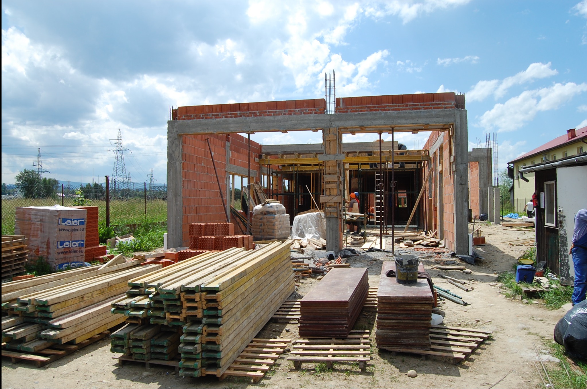 Budowa domu z czerwonej cegły, widoczne ściany w stanie surowym, drewniane stemple podtrzymujące strop, materiały budowlane na paletach, robotnicy wewnątrz konstrukcji, słupy wysokiego napięcia w tle.