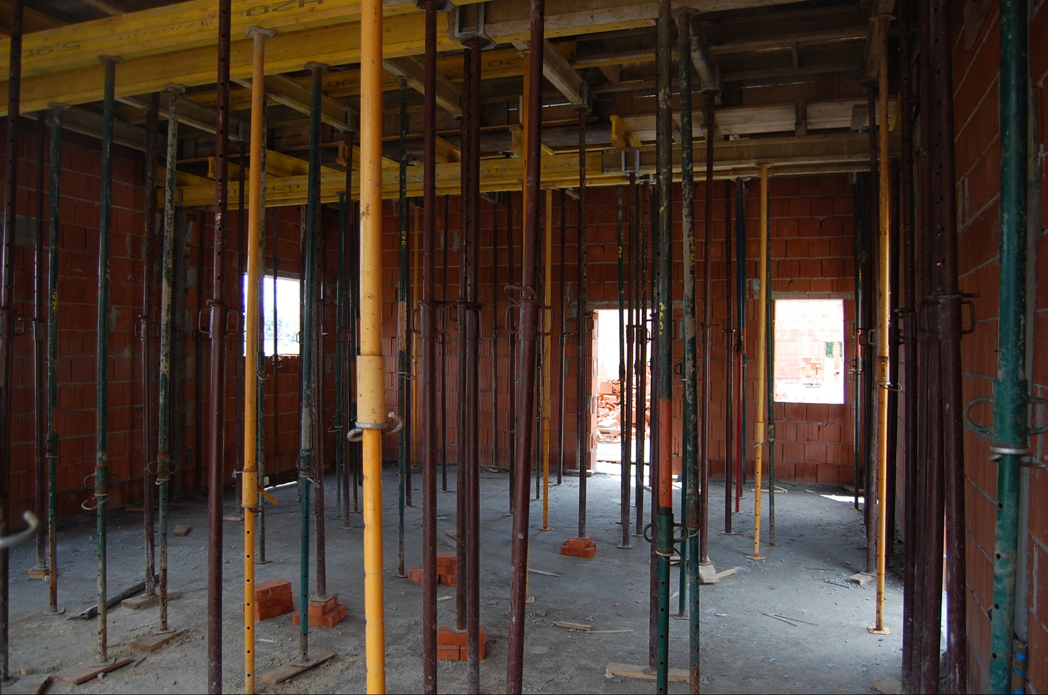 Wnętrze surowego domu w trakcie budowy: ceglane ściany, żółte i zielone stemple podtrzymujące strop, widoczne okna i drzwi w stanie surowym.