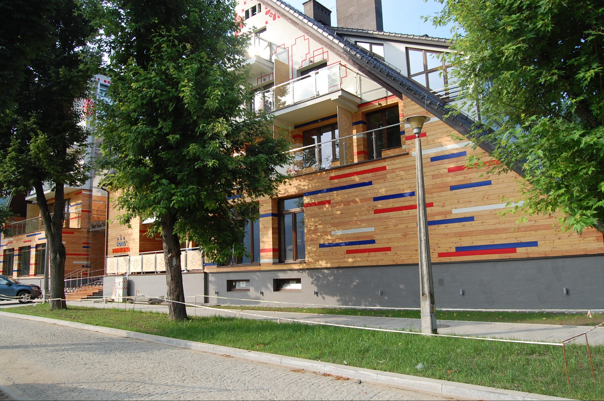 Nowoczesny budynek hotelowy z drewnianą elewacją, zdobiony poziomymi pasami w kolorach czerwonym, niebieskim i szarym, balkony z szklanymi balustradami, widok z poziomu ulicy.