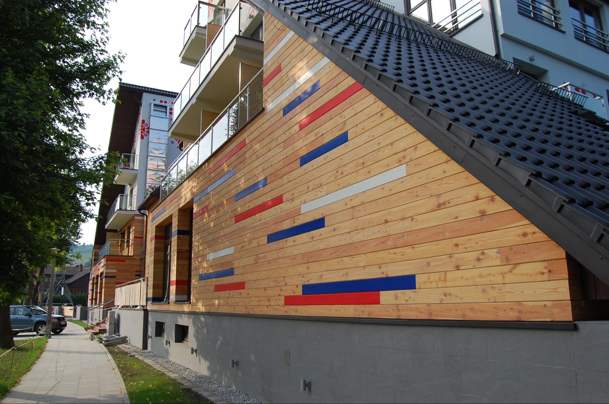 Nowoczesny budynek z drewnianą elewacją, ozdobioną poziomymi pasami w kolorach czerwonym, białym i niebieskim, widok z chodnika.