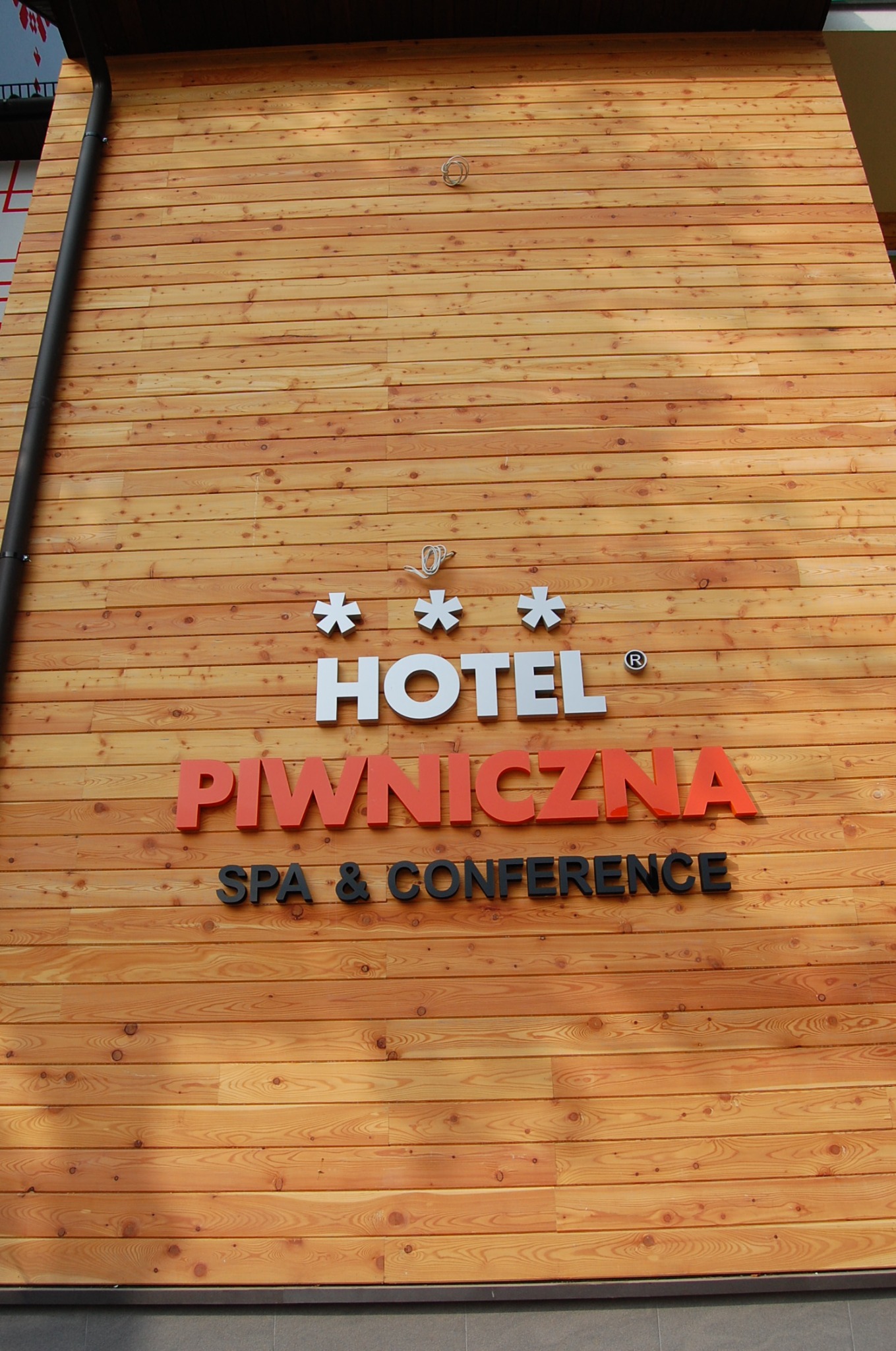 Ściana budynku obłożona jasnym drewnem z napisem 'Hotel Piwniczna Spa & Conference' w kolorach białym, pomarańczowym i czarnym.