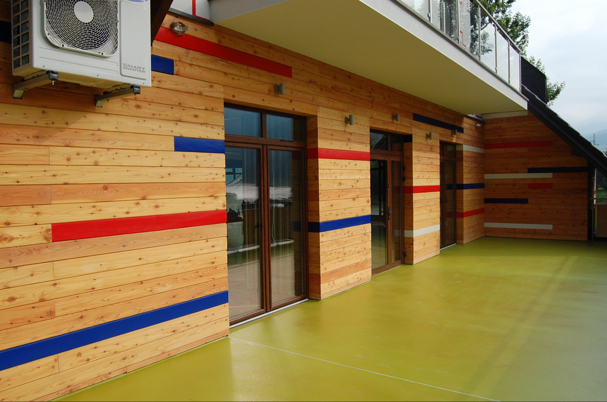 Elewacja domu z jasnego drewna z poziomymi pasami w kolorach czerwonym i niebieskim, widoczny klimatyzator, przeszklone drzwi balkonowe i zielona posadzka balkonu.