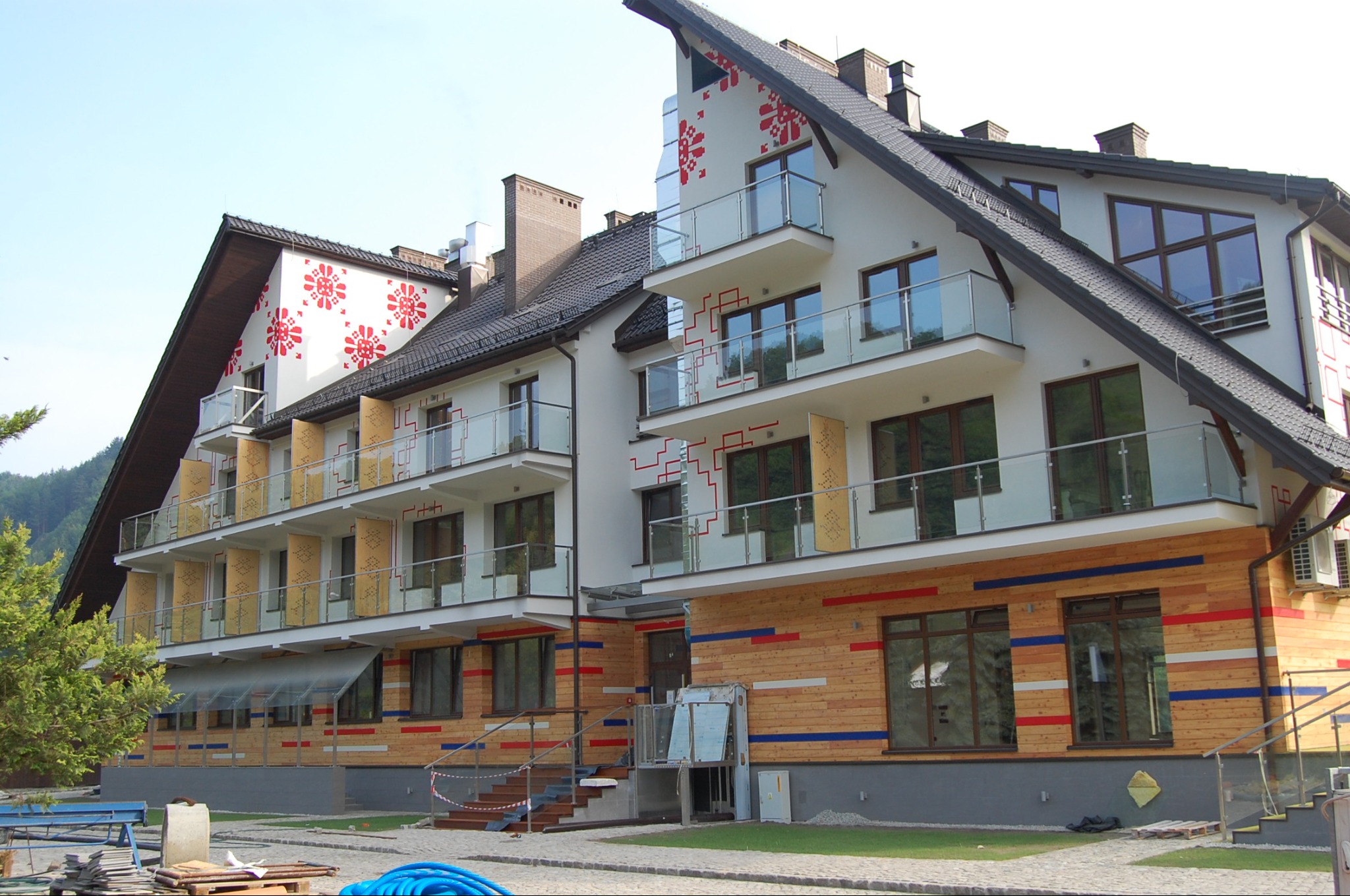 Trzykondygnacyjny budynek z białymi ścianami ozdobionymi czerwonymi wzorami kwiatowymi i drewnianą elewacją z poziomymi pasami w kolorach czerwonym, białym i niebieskim, balkony z szklanymi...