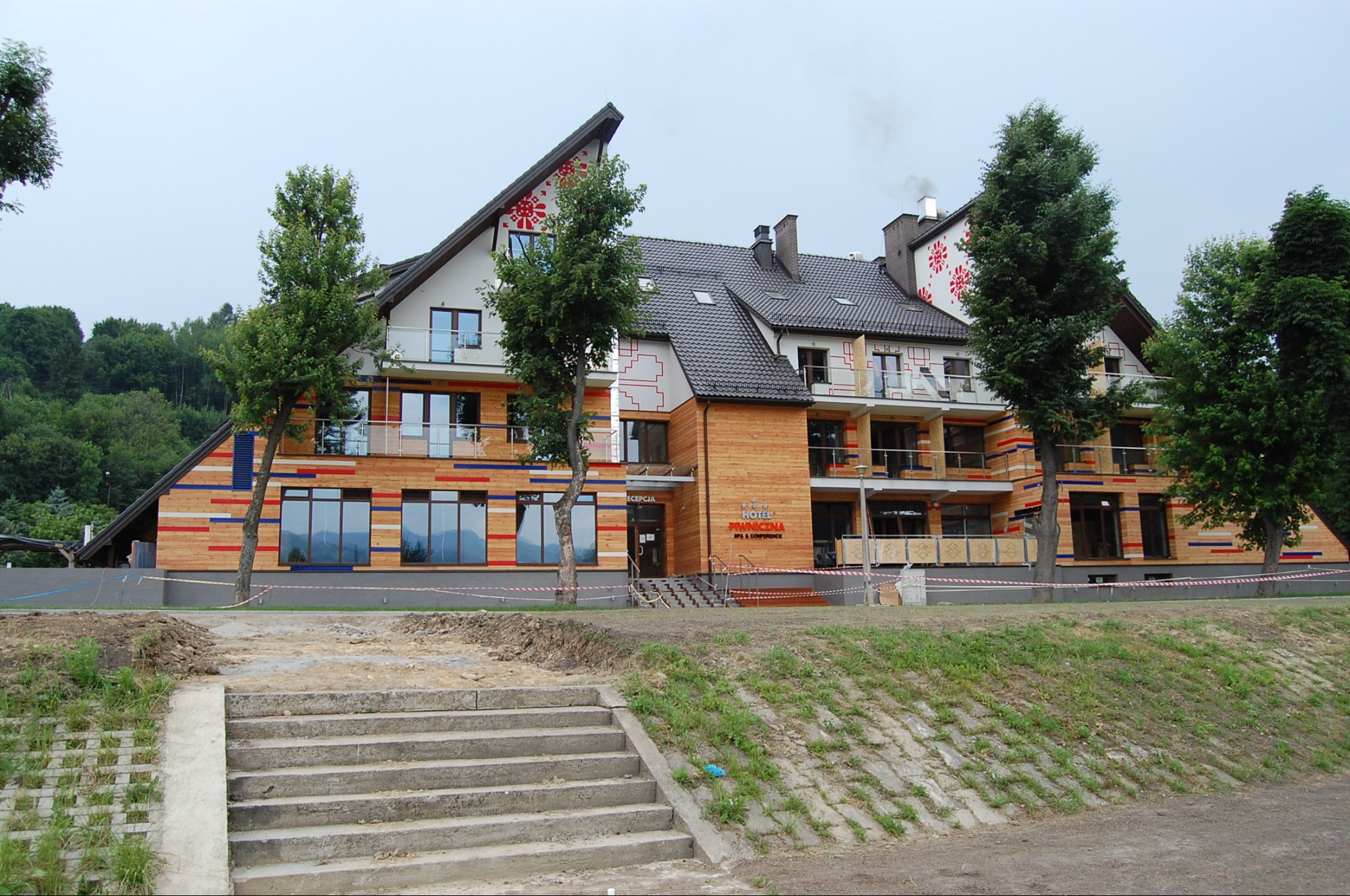 Nowoczesny budynek hotelowy z elementami drewnianej elewacji, zdobiony motywami ludowymi, widoczne schody wejściowe i teren w trakcie prac wykończeniowych.