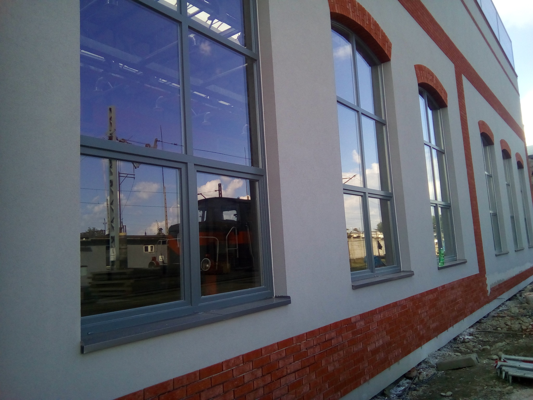 Szara elewacja budynku z ceglaną podmurówką i obramowaniami okien, w których odbija się lokomotywa i słupy trakcyjne.