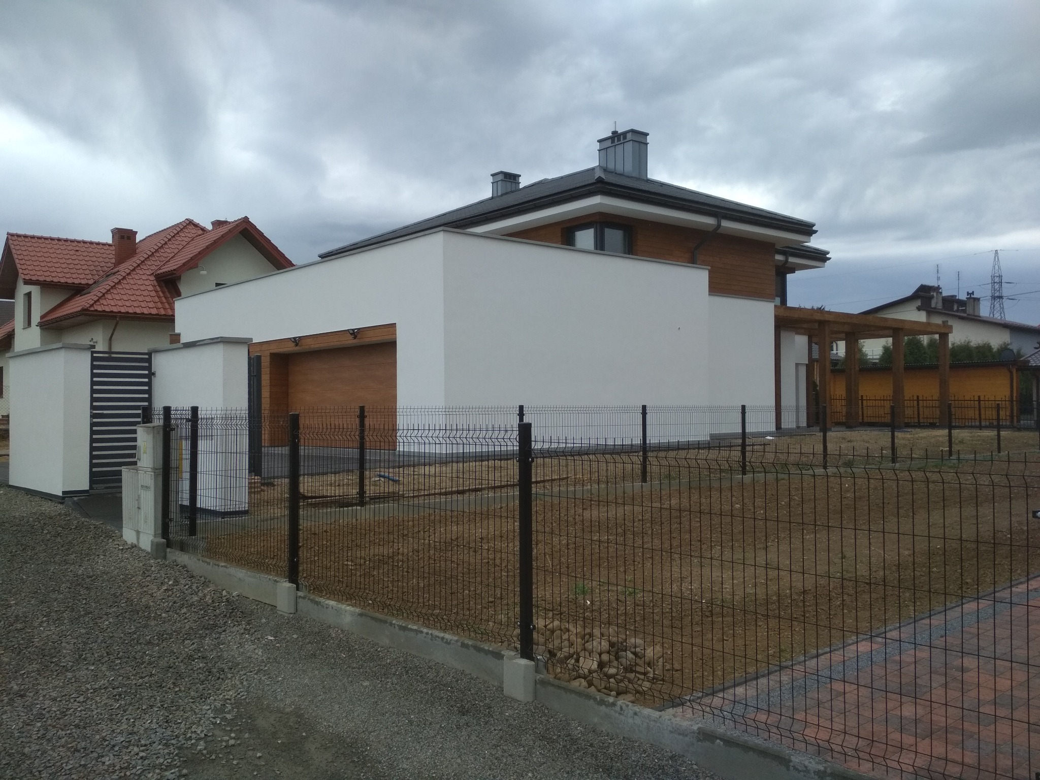 Nowoczesny dom jednorodzinny z płaskim dachem i drewnianą elewacją, widoczny zza czarnego ogrodzenia panelowego na tle pochmurnego nieba.