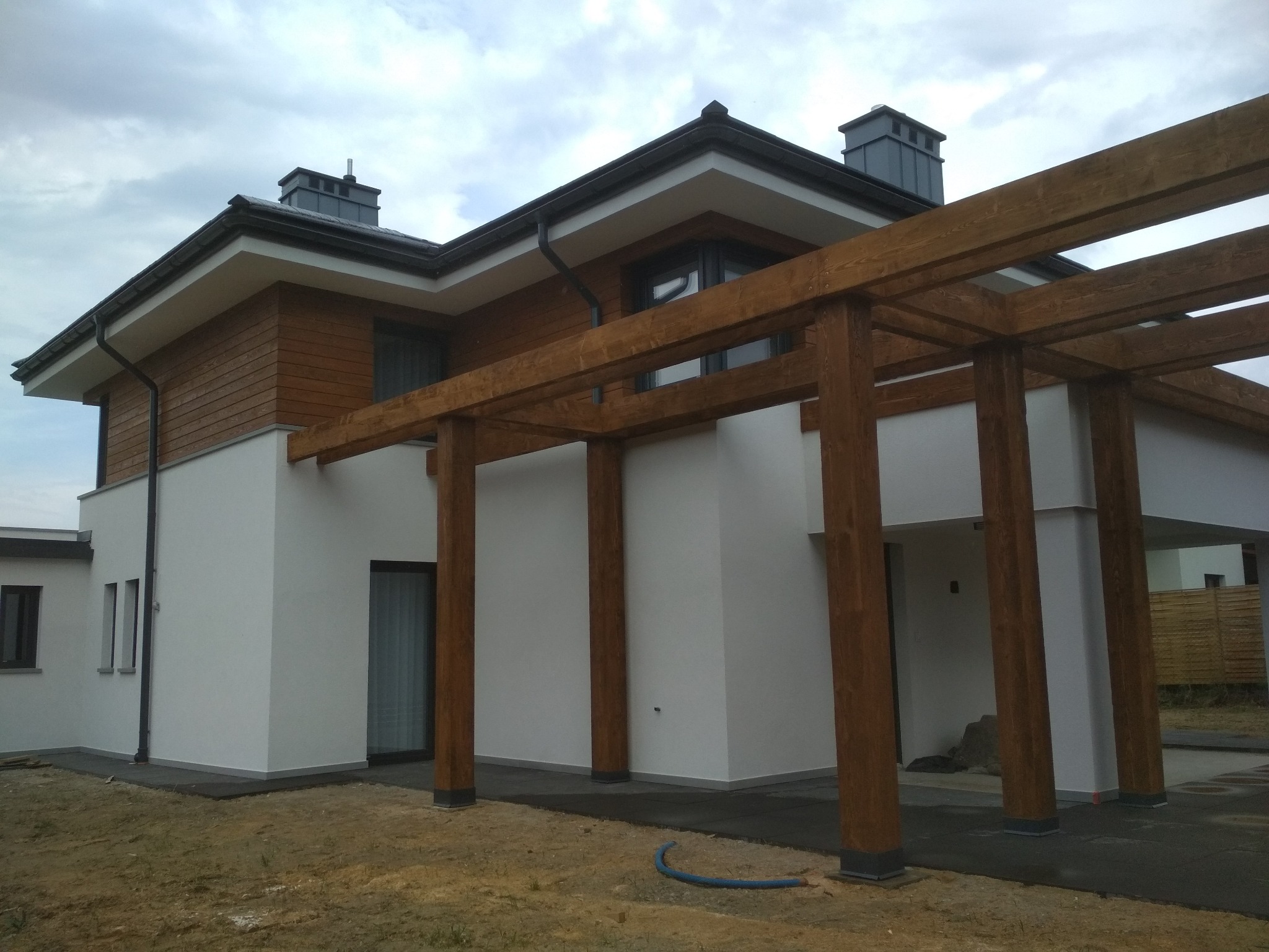 Nowoczesny dom z białą elewacją, drewnianym wykończeniem i pergolą z belek, widok z zewnątrz w pochmurny dzień.