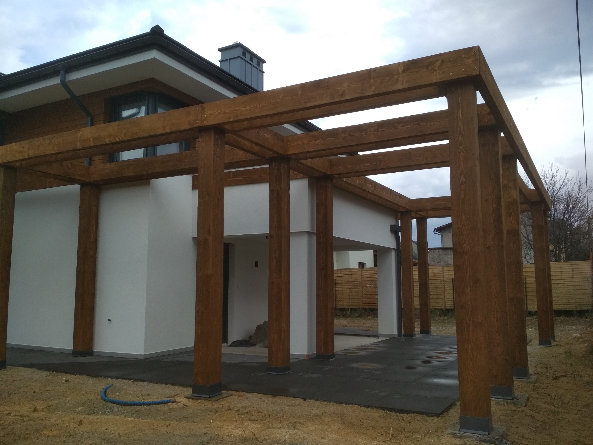 Nowoczesny dom z białymi ścianami i drewnianą konstrukcją pergoli w kolorze orzechowym, z szarą podłogą z płyt i niebieskim wężem na ziemi.