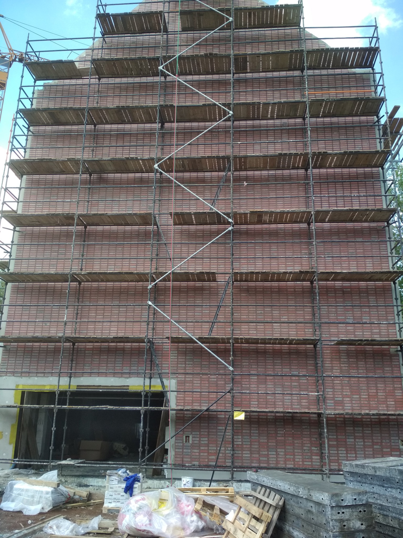 Wznoszony budynek z czerwonej cegły, otoczony rusztowaniem, widoczne materiały budowlane i palety na pierwszym planie, fragment dźwigu w tle.