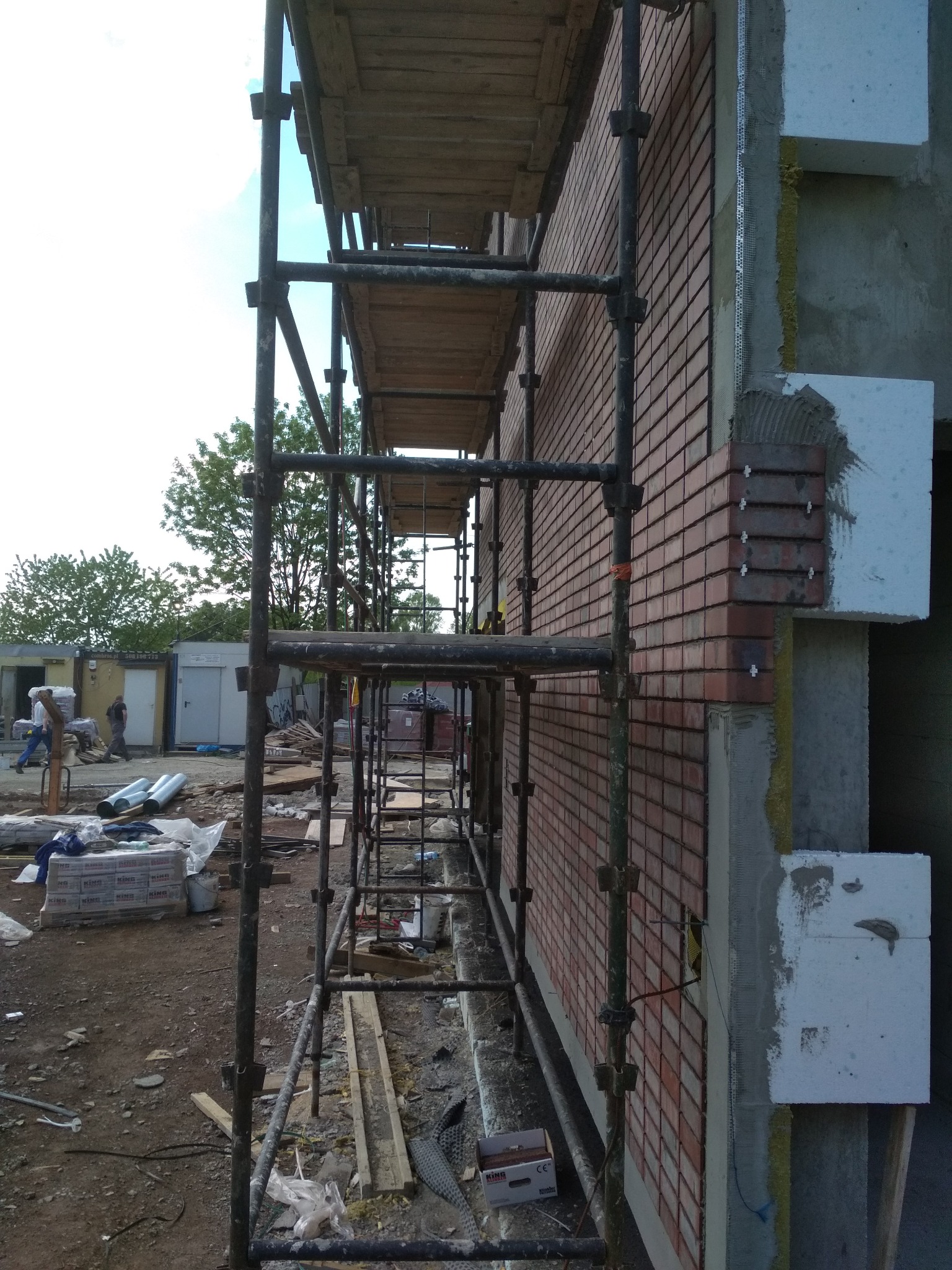 Budowa domu: fragment elewacji z cegły klinkierowej na rusztowaniu, widoczne ocieplenie styropianem i elementy montażowe, w tle teren budowy.